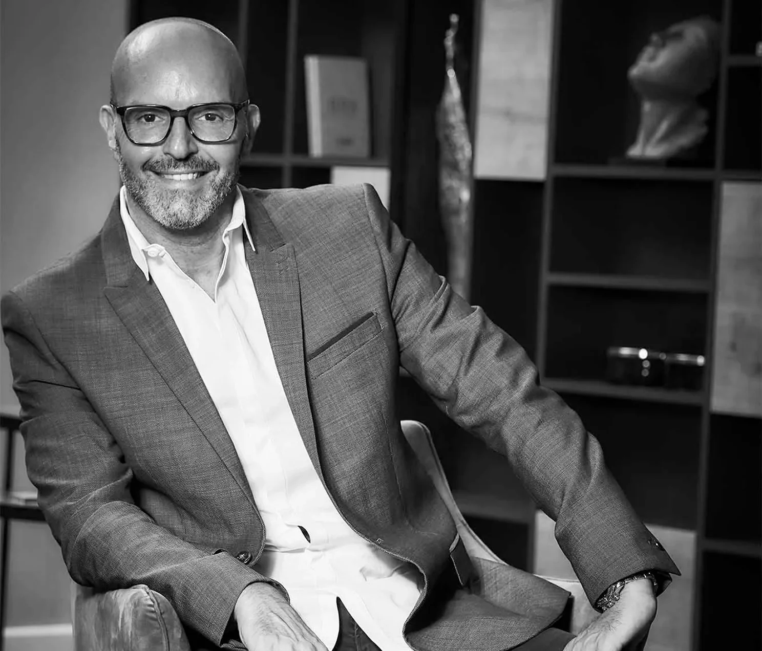 Professional black-and-white portrait of man wearing glasses and a blazer, seated and smiling in a modern office setting.