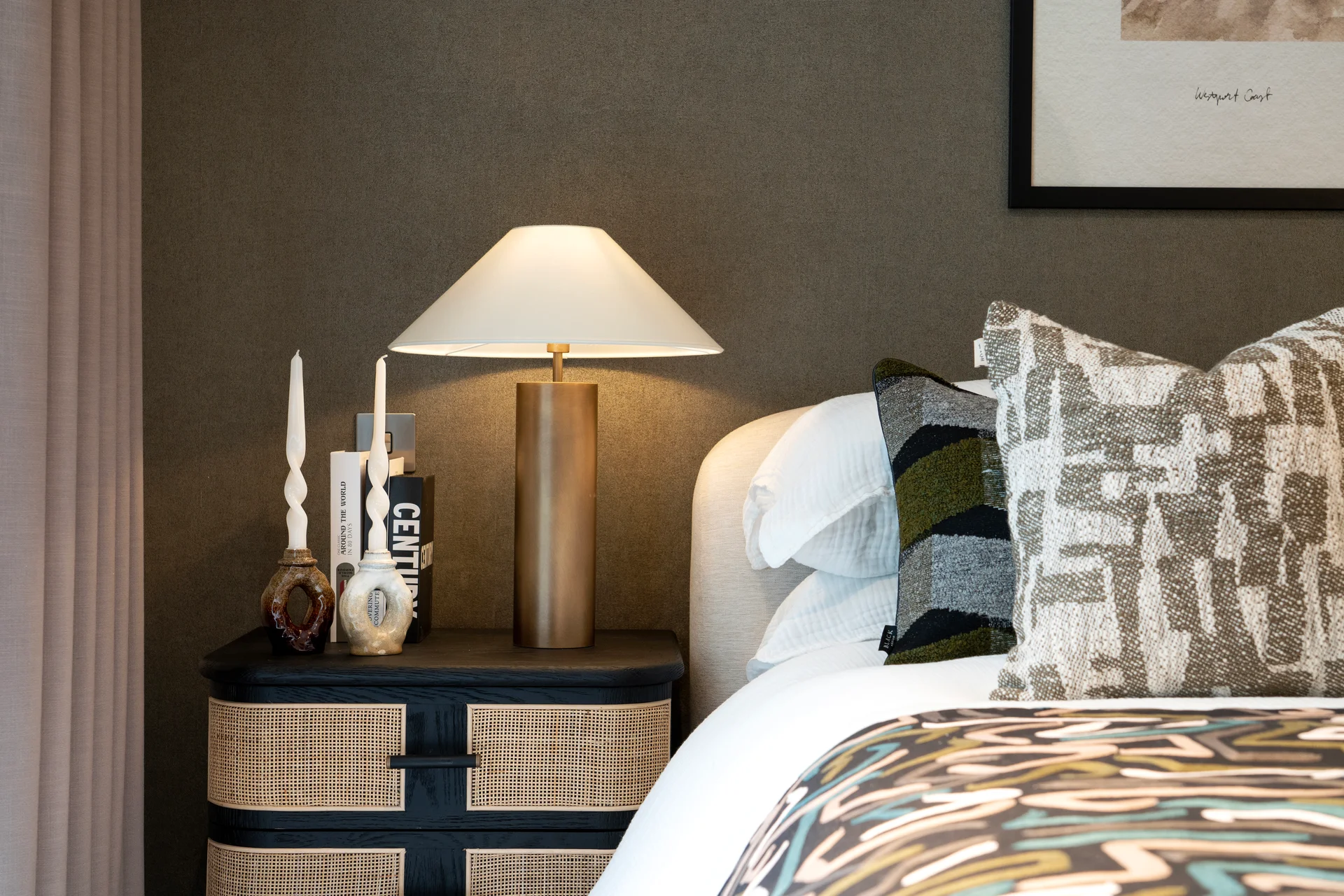 Close-up of a bedroom featuring a rattan-fronted bedside unit, brass lamp, sculptural candlesticks, and patterned bedding with textured cushions.