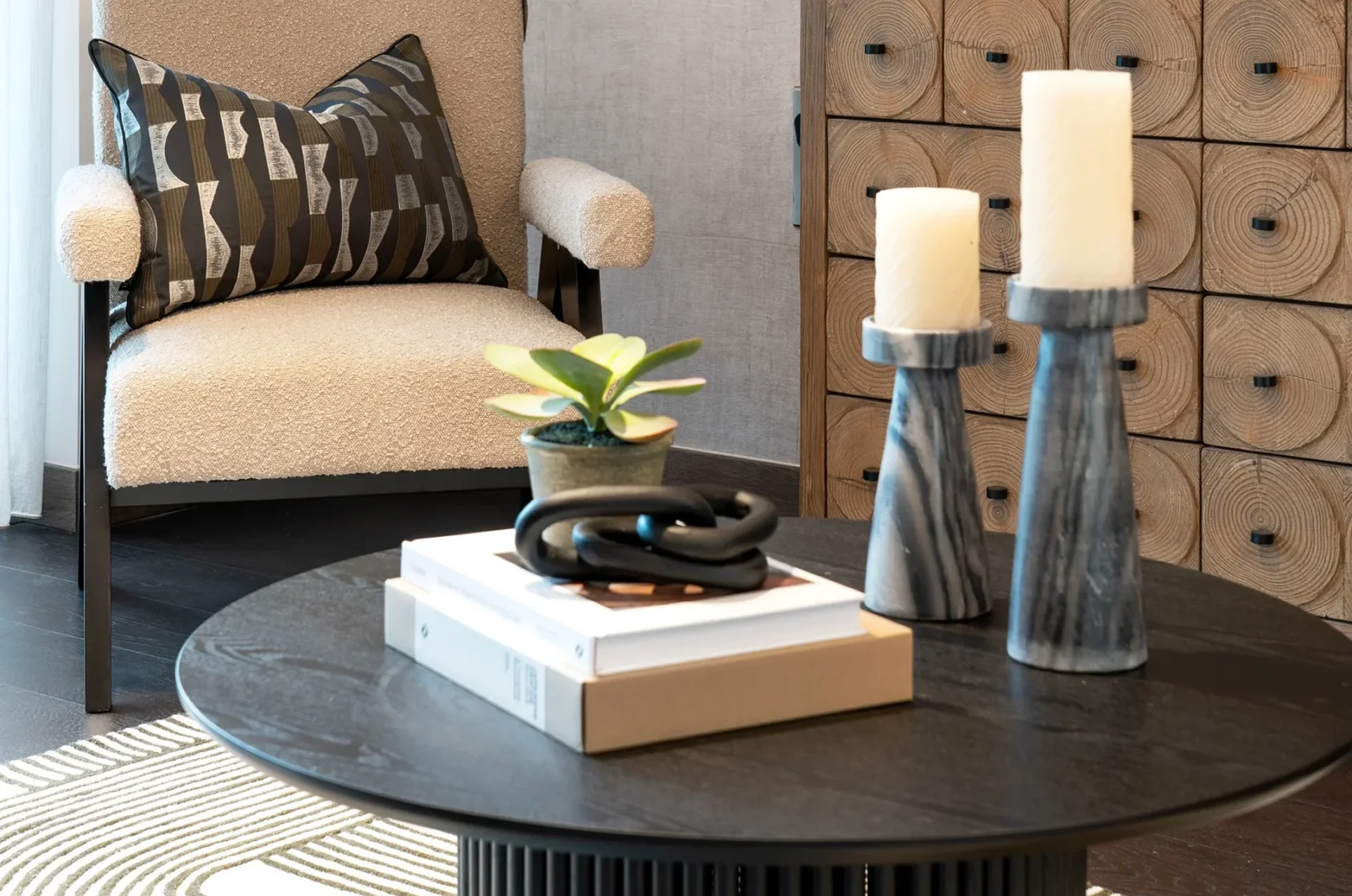 Modern lounge corner with boucle armchair, decorative wooden cabinet, black round coffee table, sculptural candles, and pampas grass in a vase.