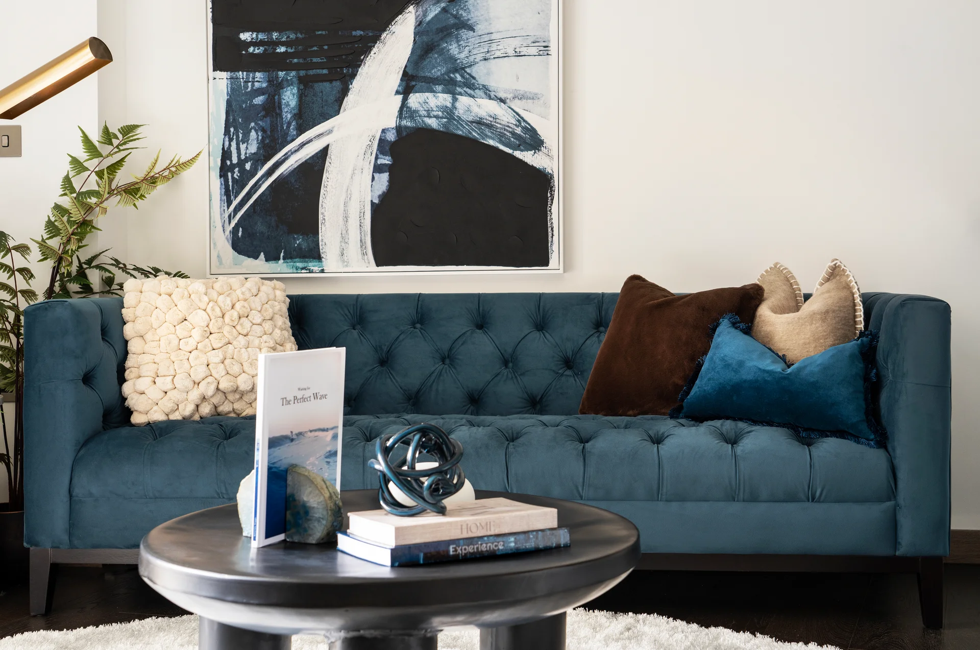 Living room with a deep blue velvet sofa, mixed textured cushions, large abstract wall art, and a round black coffee table with decor books.