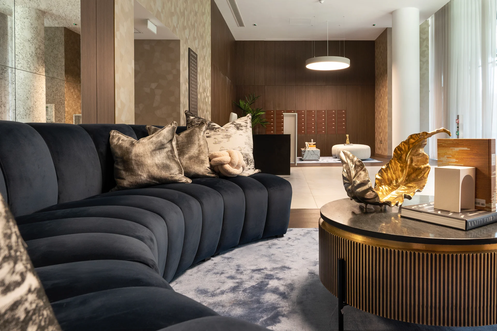 Close-up view of a curved navy velvet sofa with plush cushions, a round coffee table with gold decor, and a modern, well-lit apartment lobby in the background.