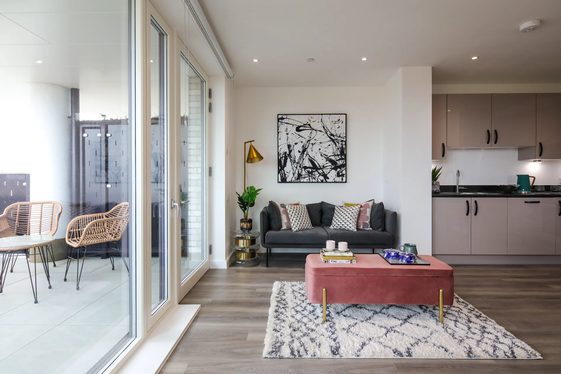 Bright and compact open-plan living space with a grey sofa, modern art, soft furnishings, and a pink ottoman coffee table, leading to a balcony with wicker chairs.