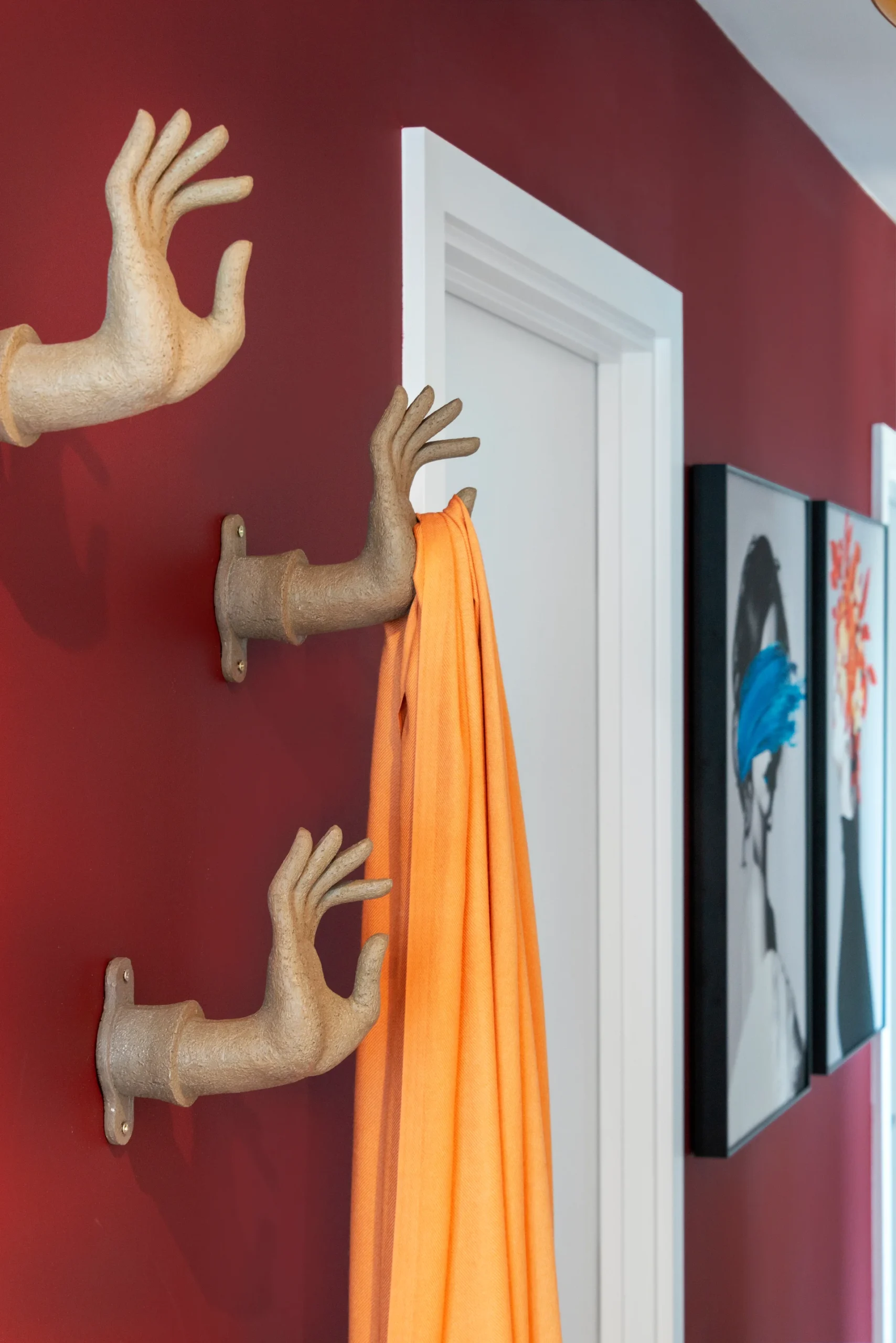 Two wall-mounted hand sculptures hold an orange fabric, with a door and colorful artwork in the background.