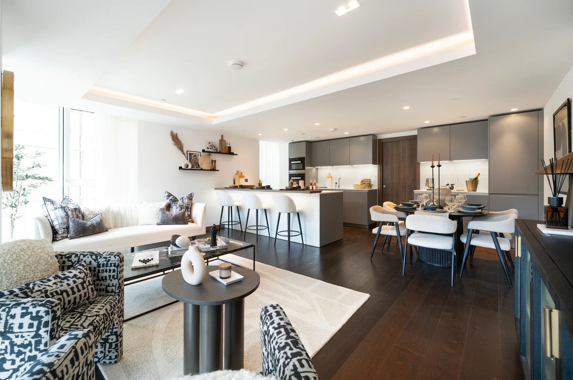 Modern living room and kitchen combo with stylish furniture, wooden flooring, and natural light from large windows.