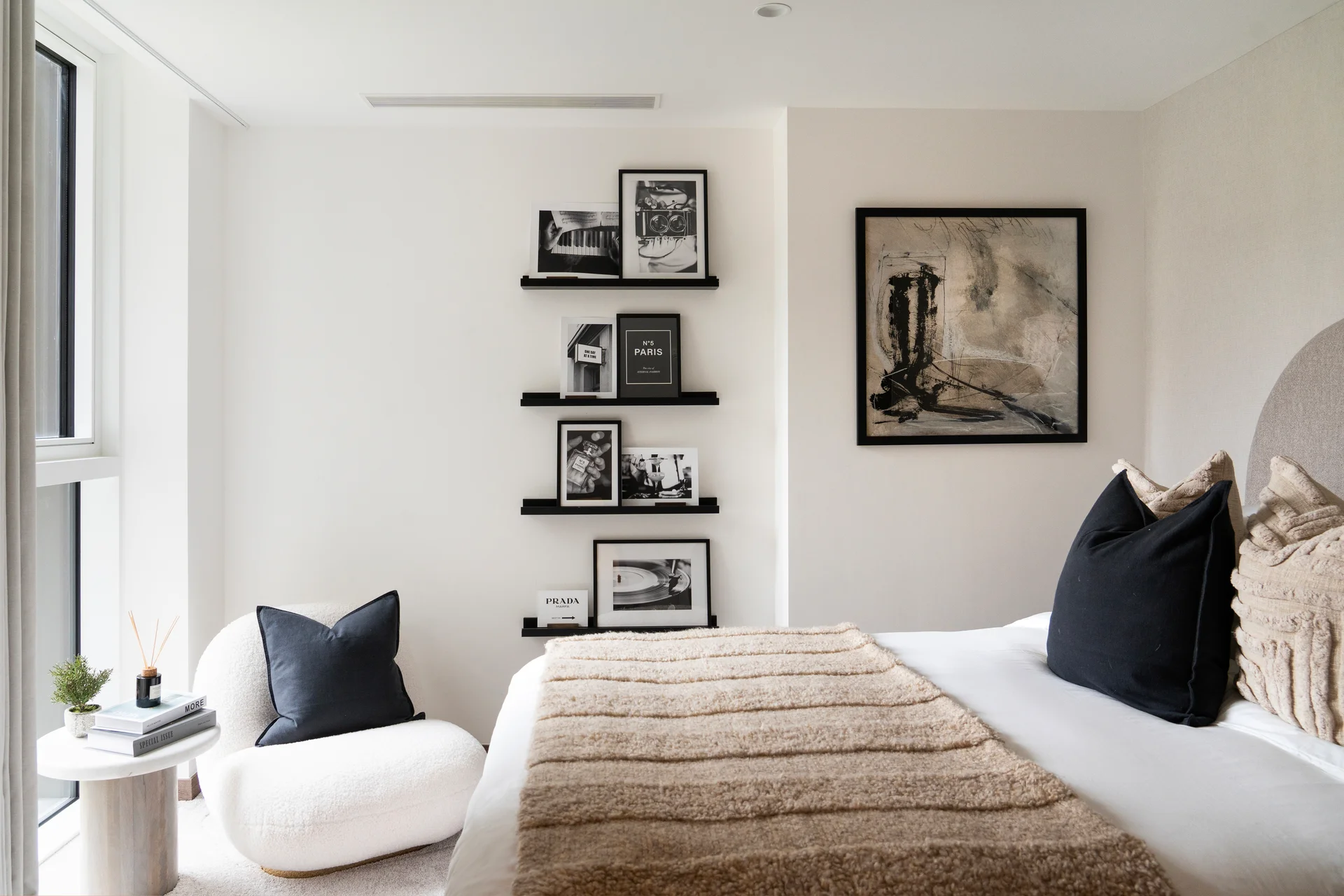 Modern bedroom featuring a neatly made bed, decorative pillows, wall art, and shelves with framed photos.