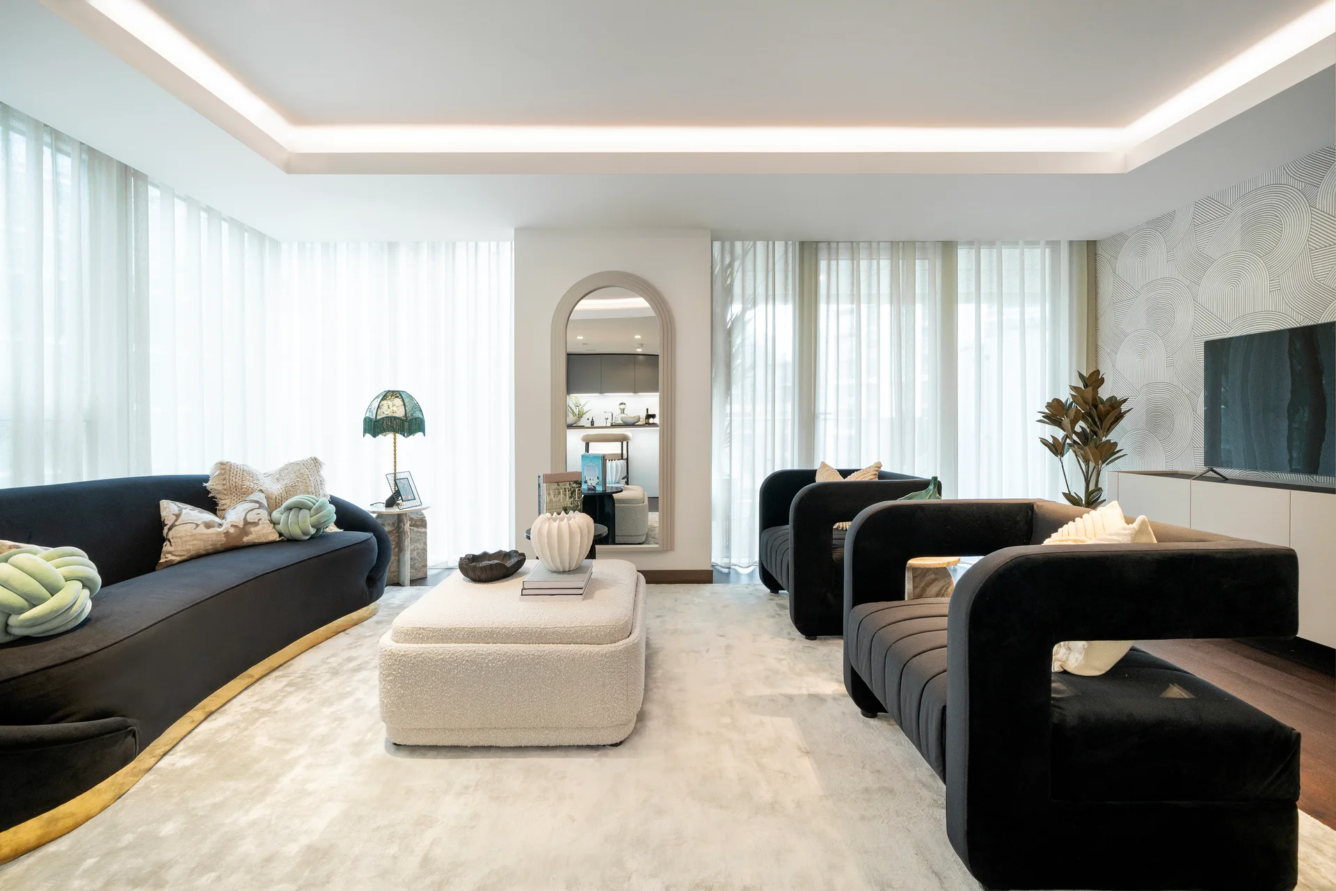 Modern living room featuring black sofas, a white coffee table, large windows with sheer curtains, and a TV unit.
