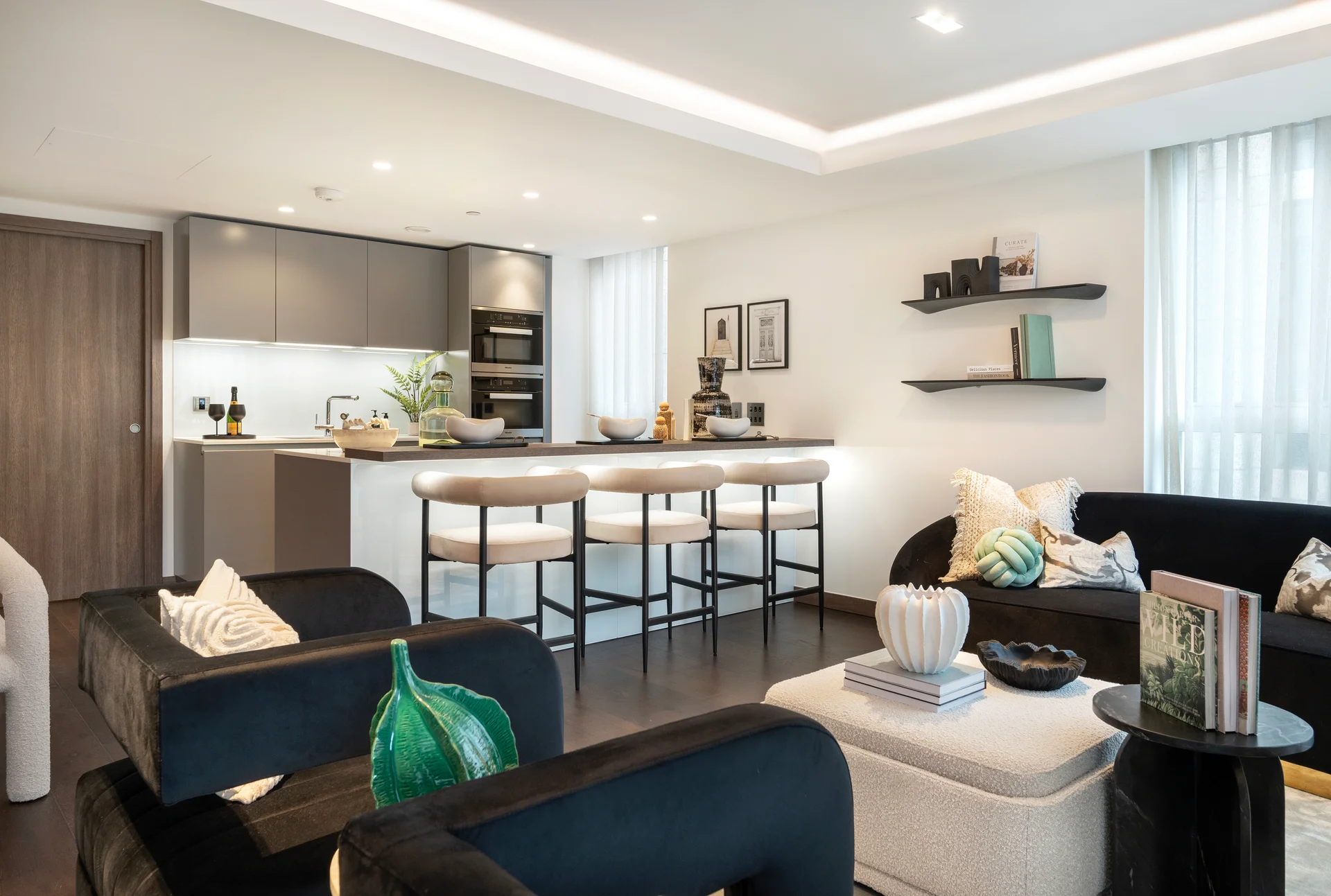 Modern living room with black sofas, a kitchen area, and decorative shelves. Bright natural light from windows.