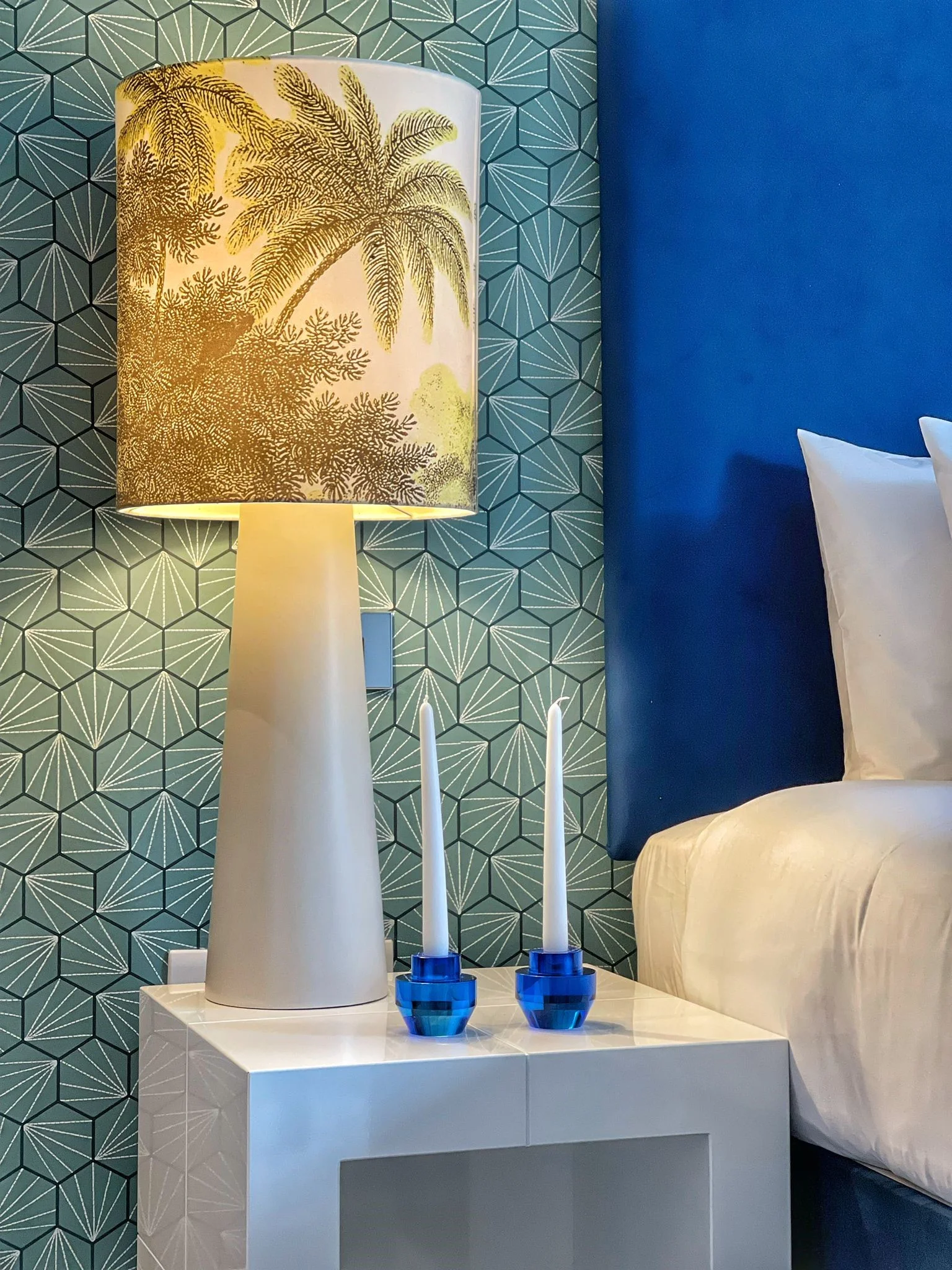 Table lamp with palm tree design and two blue candle holders on a bedside table against a textured green and blue wall.