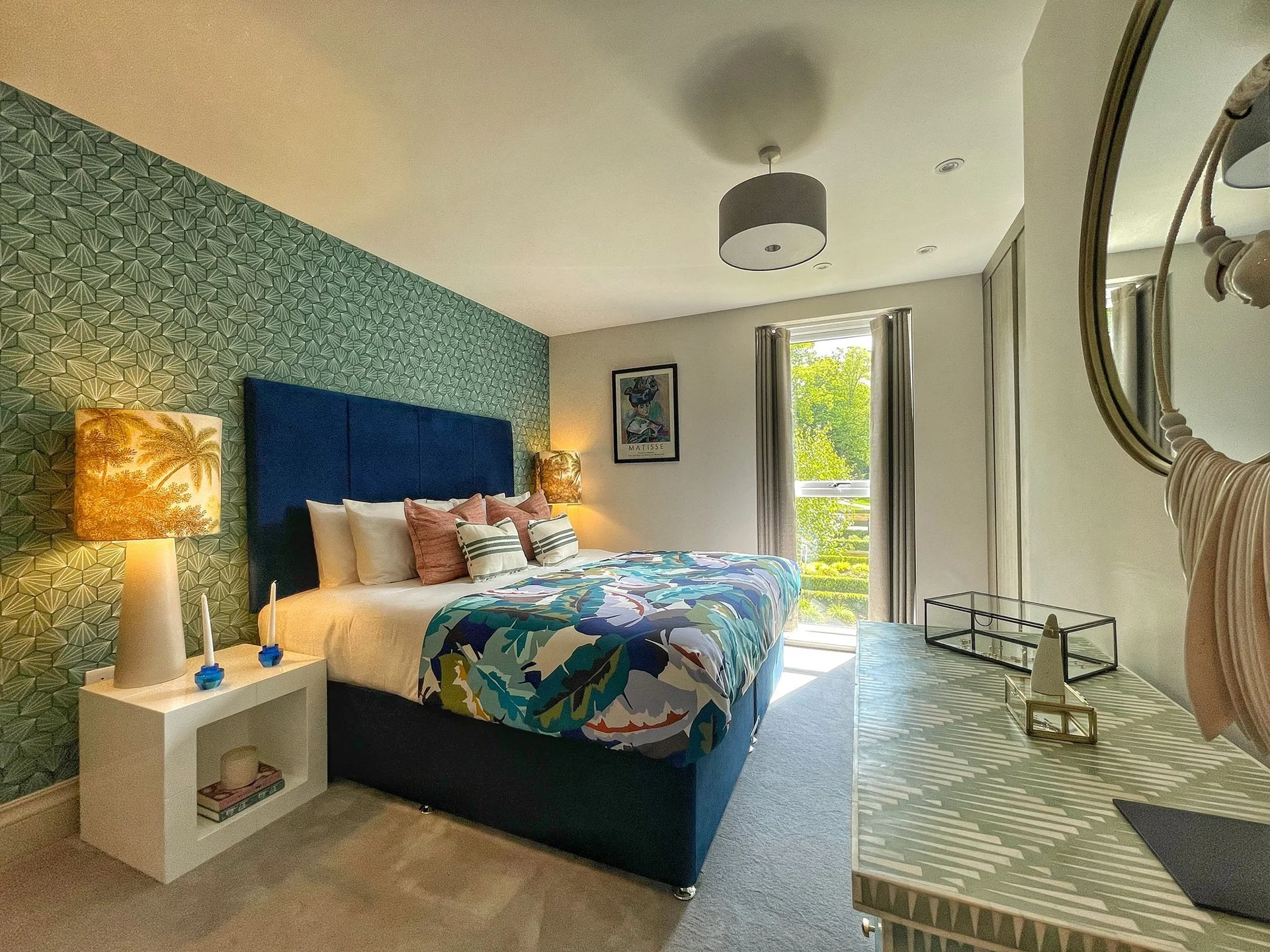 Stylish bedroom with a blue upholstered bed, colorful bedding, a decorative lamp, and large windows letting in natural light.