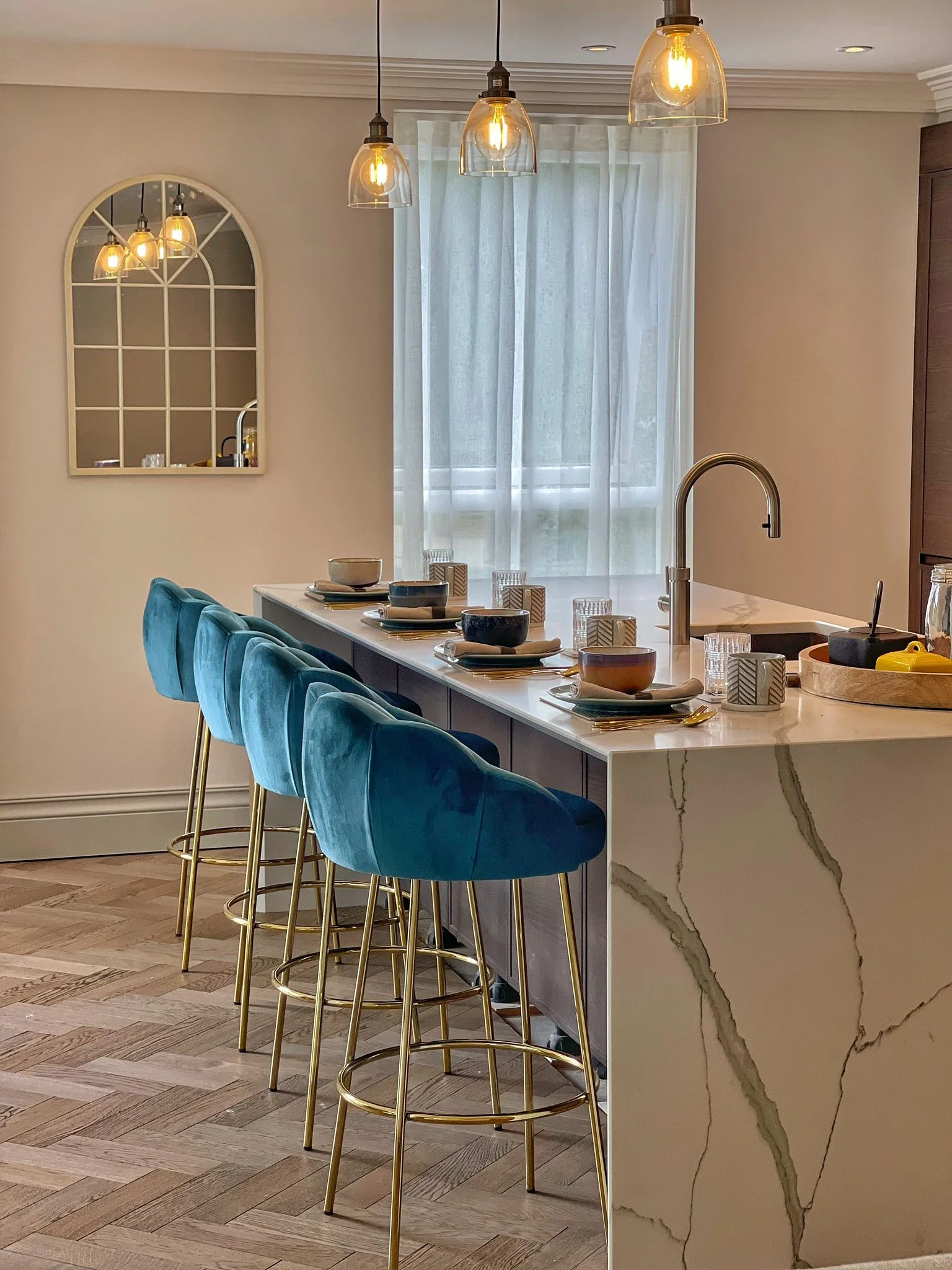 Modern kitchen with a marble island, teal bar stools, and a large window with sheer curtains. Decorative mirror on the wall.