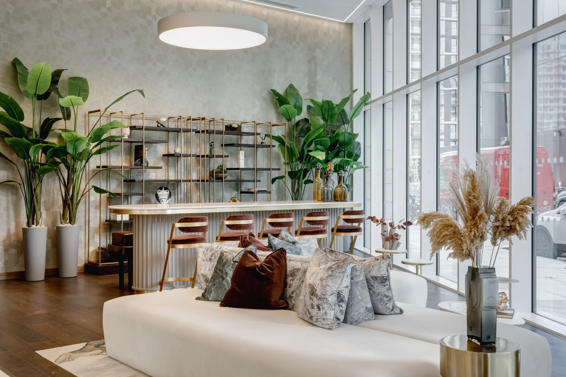 Modern reception area featuring a white sofa, decorative pillows, and large windows with greenery and a stylish bar.