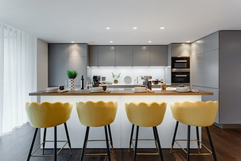Modern kitchen with sleek gray cabinets, white countertops, and yellow bar stools. Brightly lit with warm ambiance.