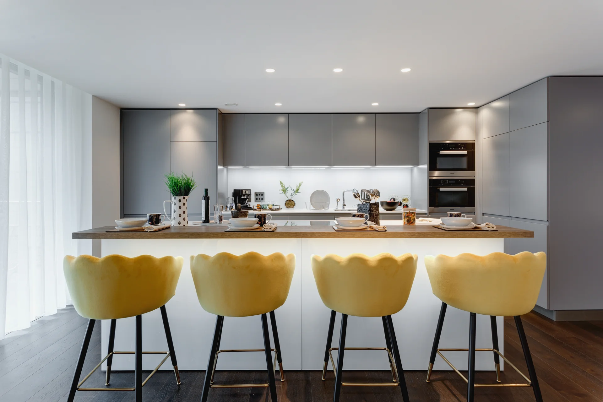Modern kitchen with sleek gray cabinets, white countertops, and yellow bar stools. Brightly lit with warm ambiance.