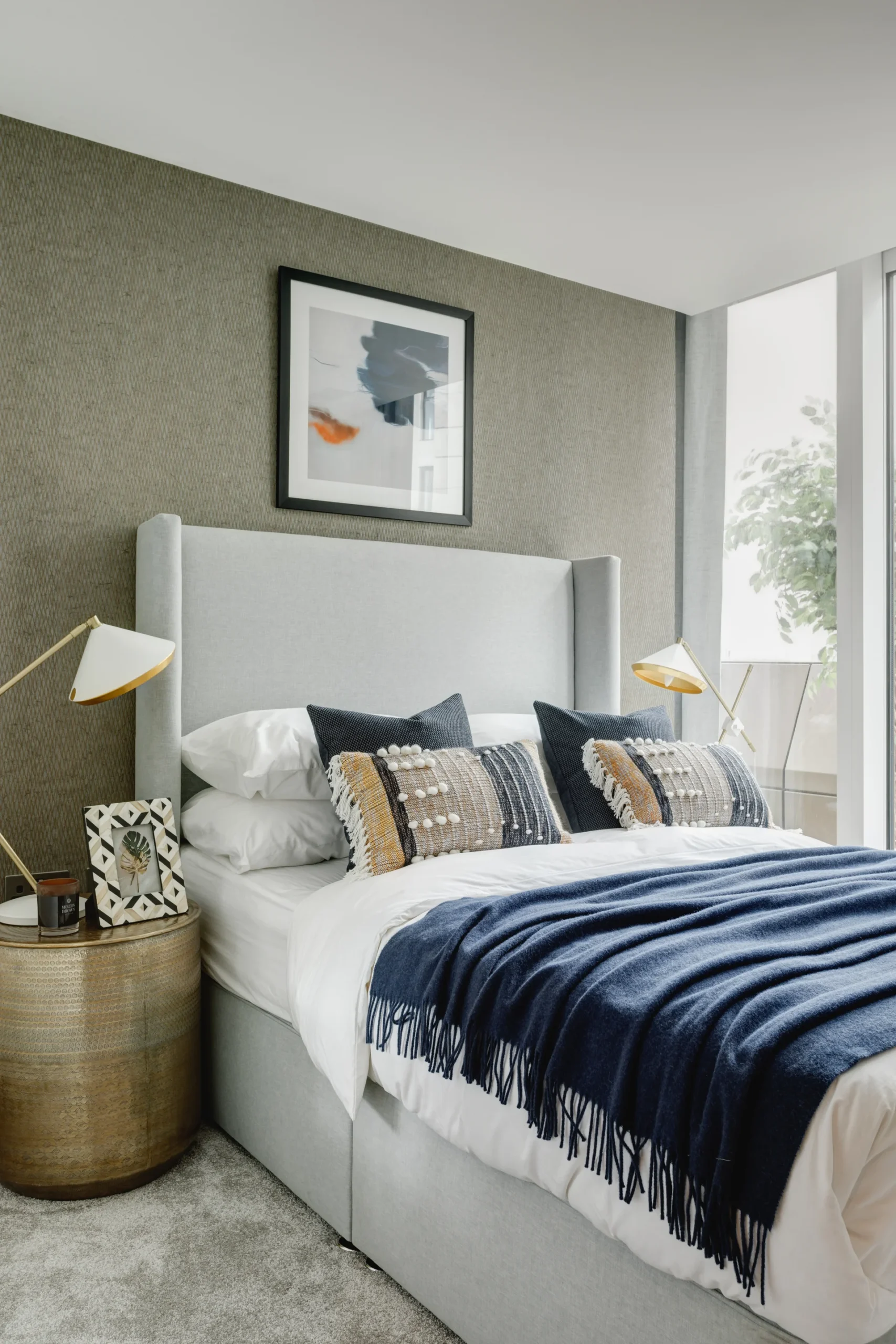 Modern bedroom featuring a gray upholstered bed, navy blanket, decorative pillows, wooden side table, and artwork.