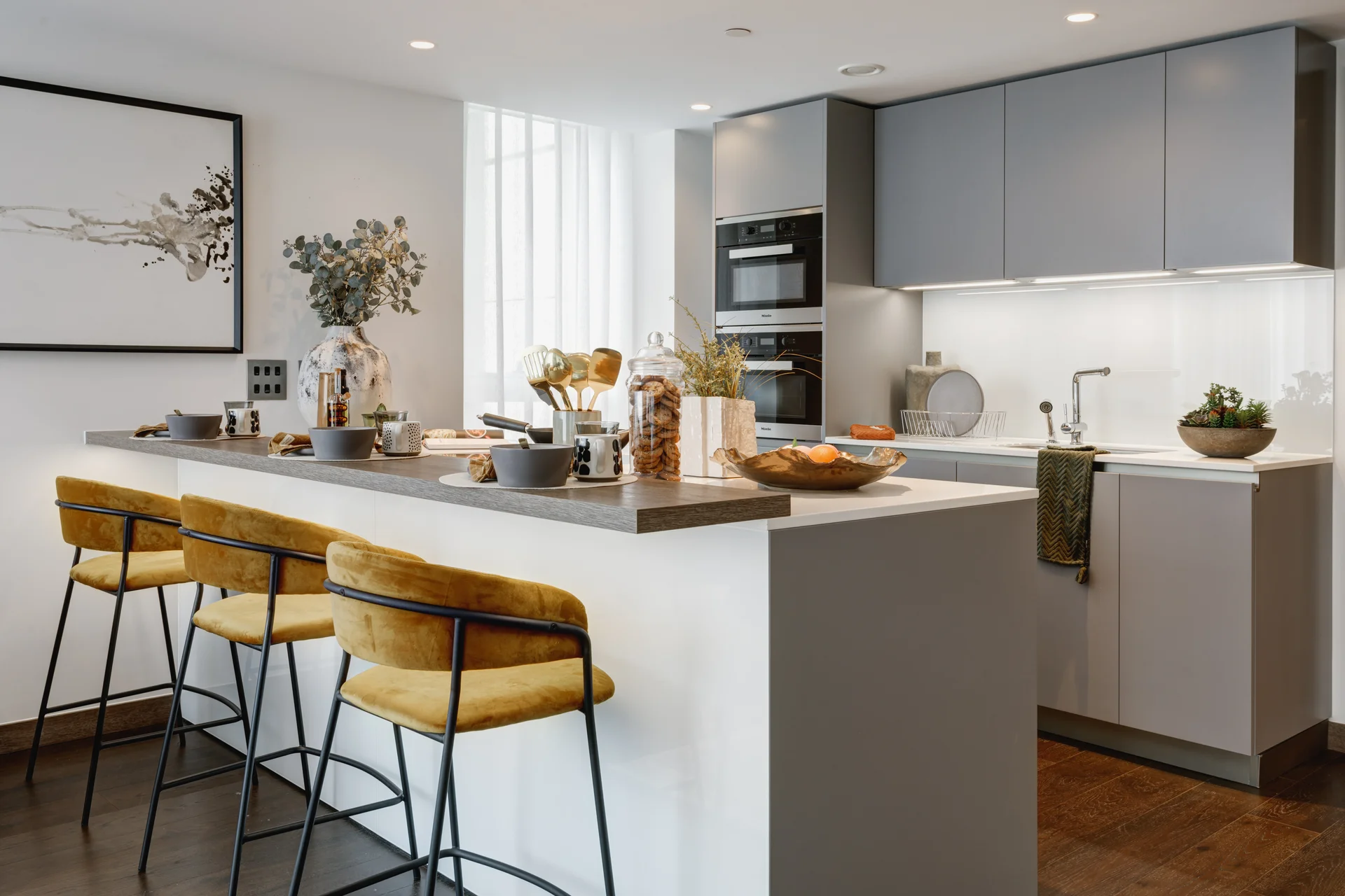 Modern kitchen with gray cabinetry, breakfast bar, yellow stools, and decorative plants on the counter.