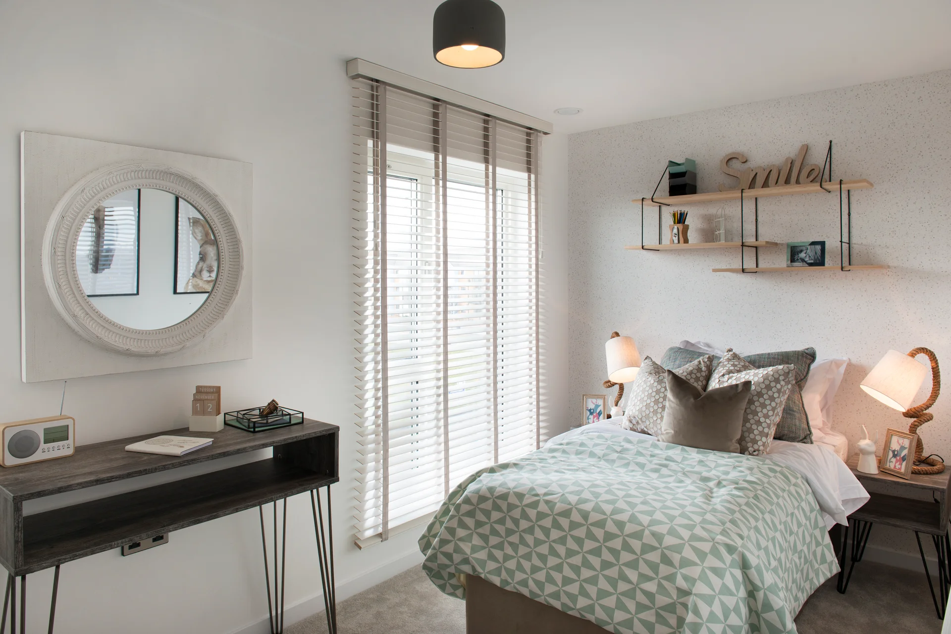 Cozy bedroom featuring a bed with a patterned blanket, a desk, shelves, and large windows with blinds.