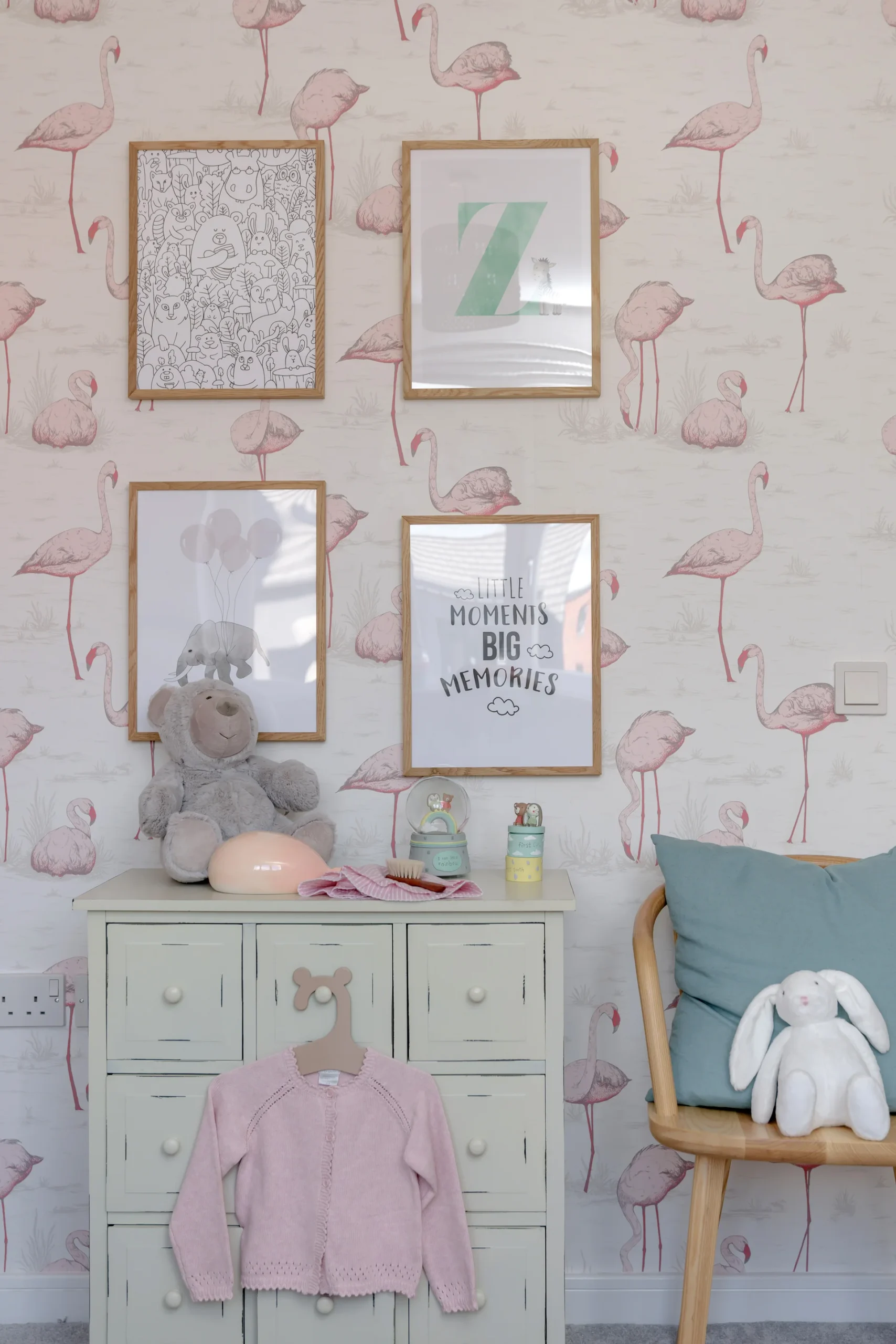 Pastel-themed nursery featuring pink flamingo wallpaper, framed art, a teal cushion, and stuffed animals on a dresser.