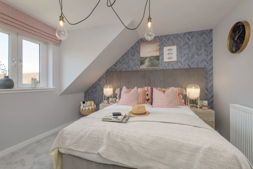 Cozy bedroom with a gray bed, pink accents, two bedside lamps, and wall art, featuring slanted ceiling and natural light.
