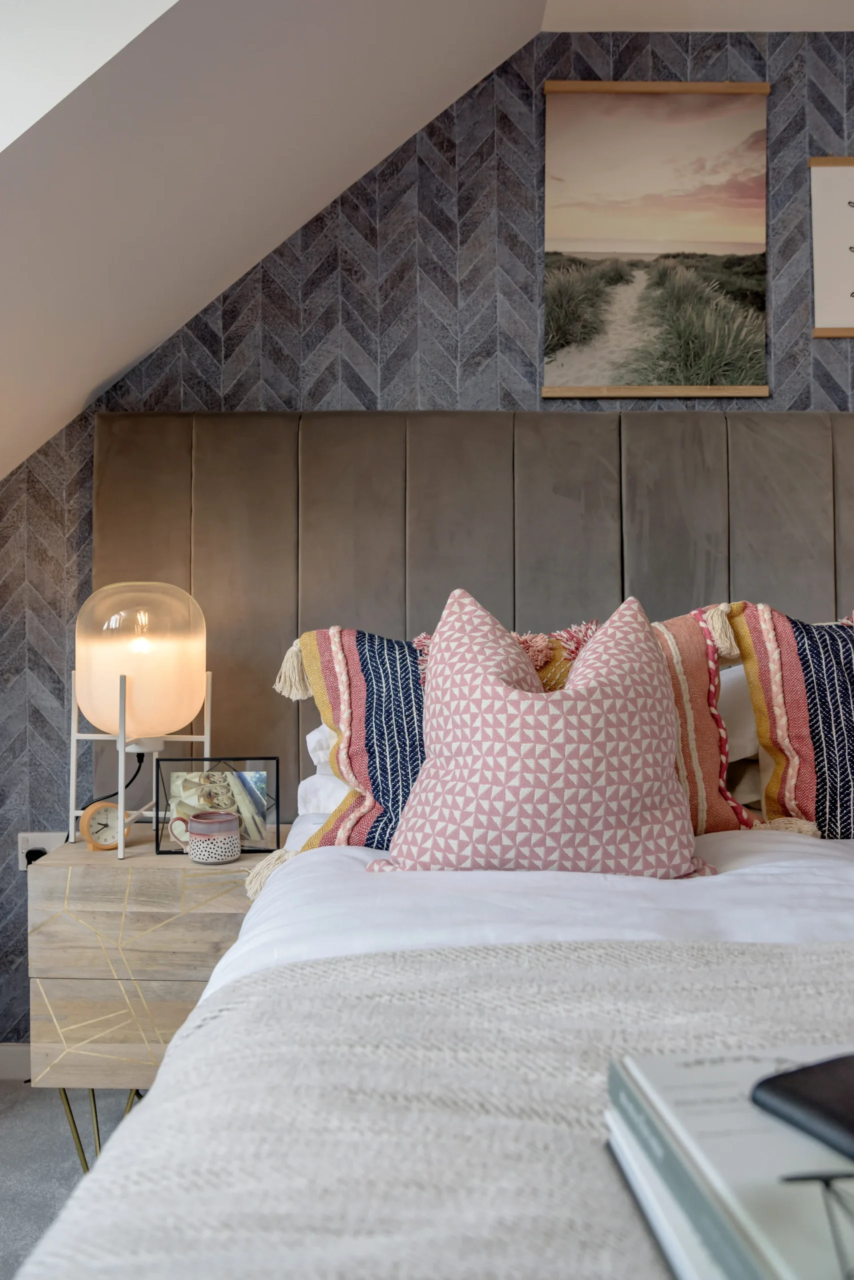 Cozy bedroom with a textured grey headboard, colorful pillows, bedside lamp, and framed artwork on the walls.