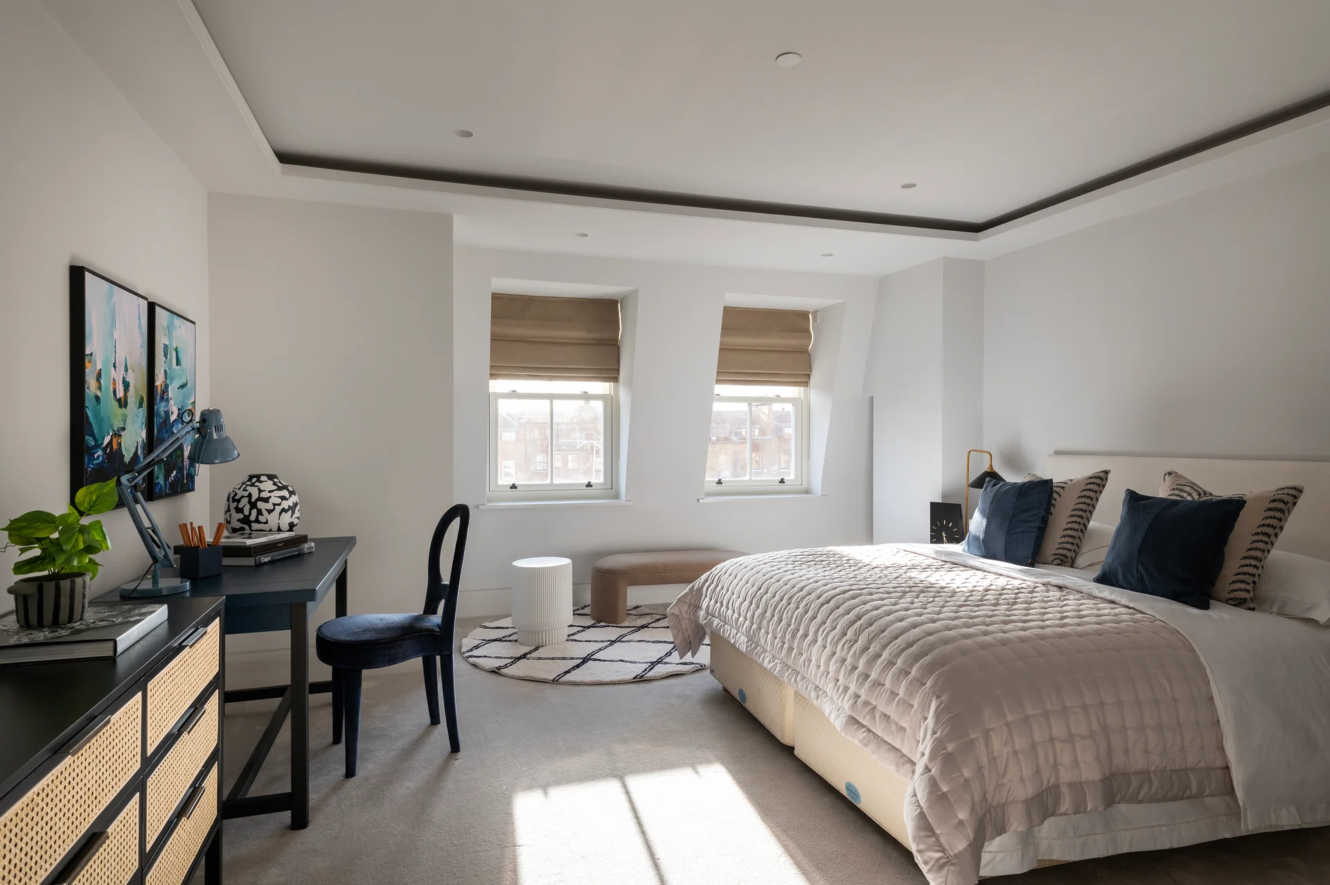 Bright, contemporary bedroom featuring a large bed, desk, chair, and natural light from windows with shades.
