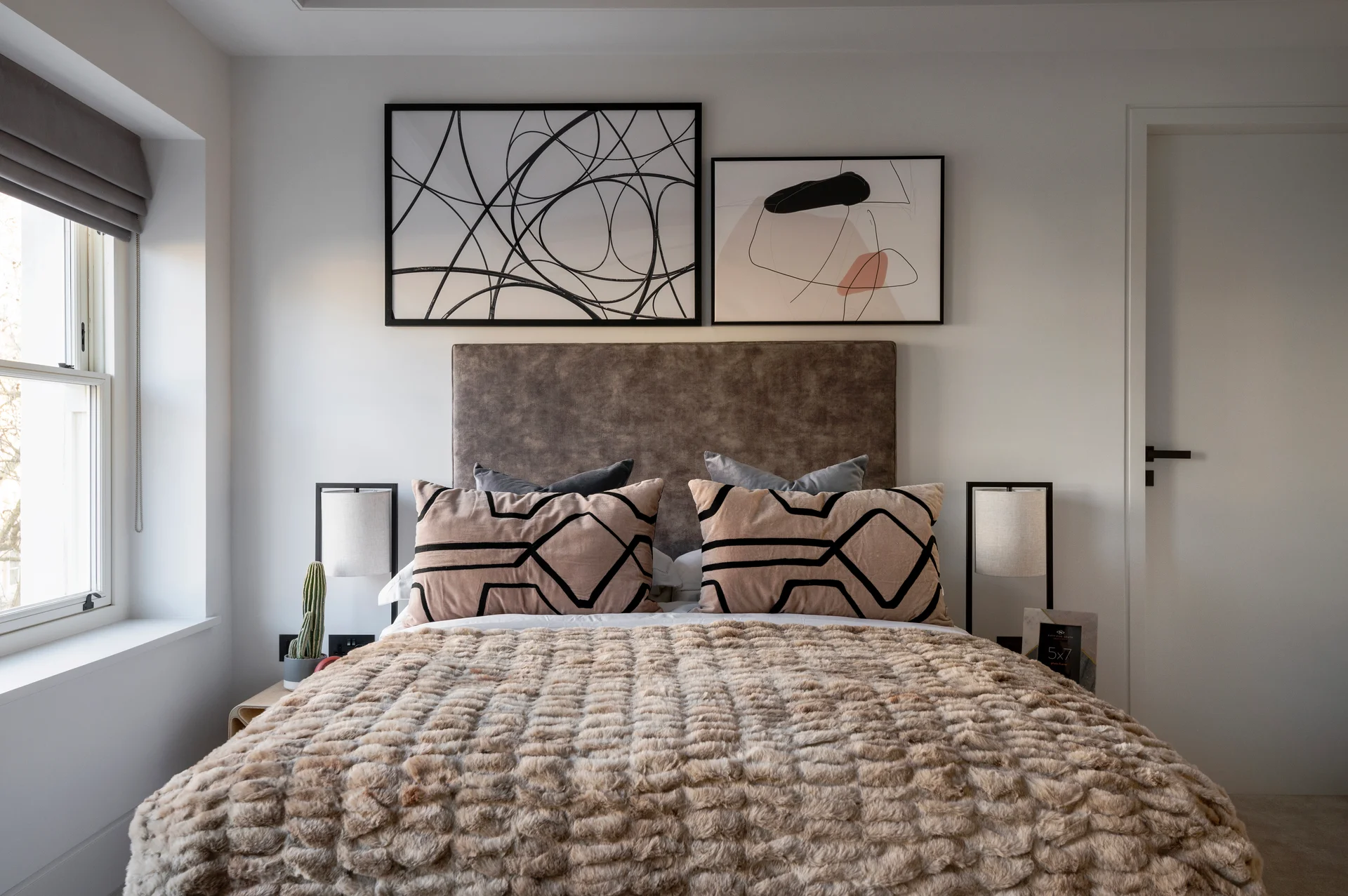 Stylish bedroom with gray upholstered headboard, patterned pillows, abstract art on walls, and natural light from the window.