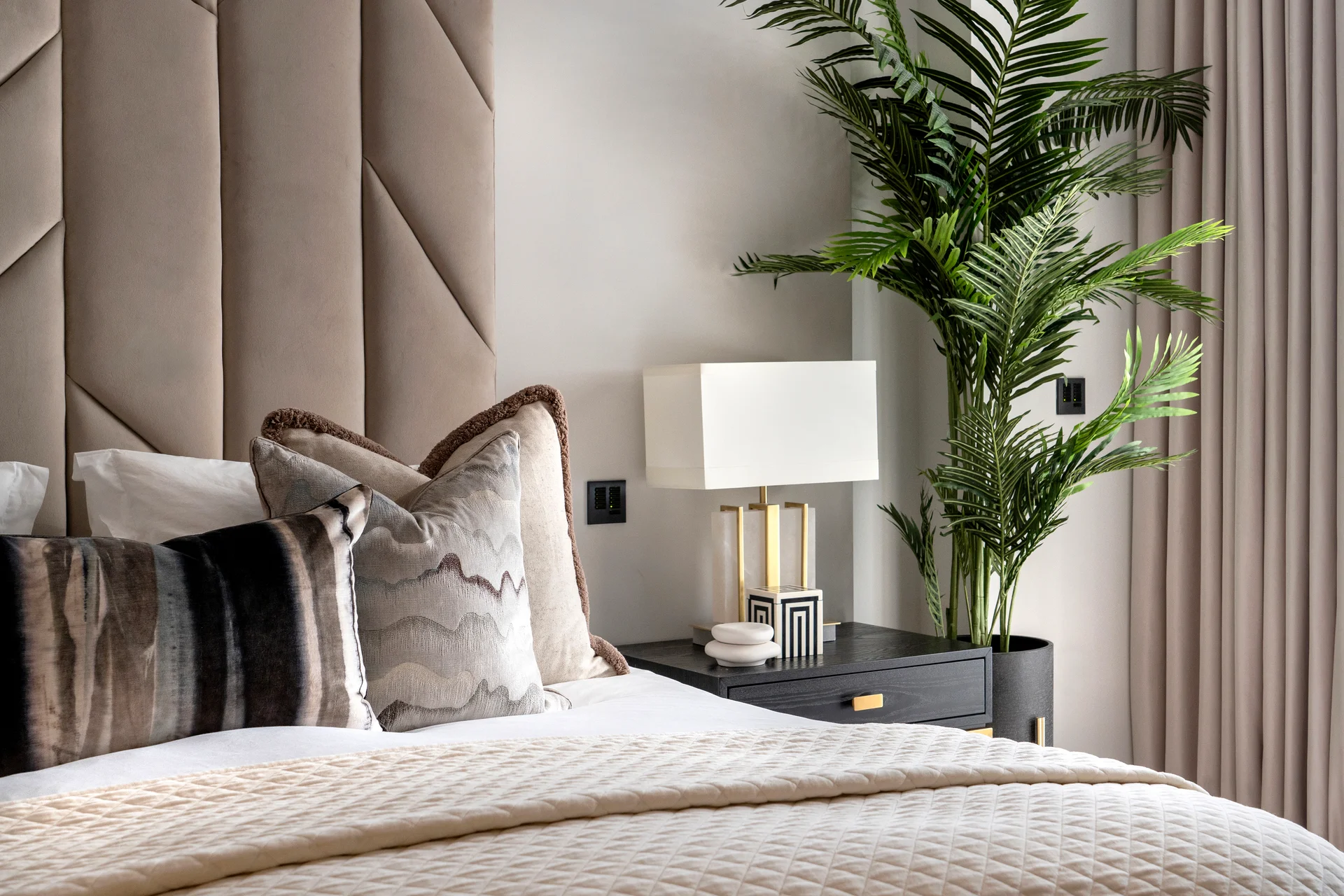 Stylish bedroom featuring a plush headboard, decorative pillows, bedside lamp, and a potted plant.