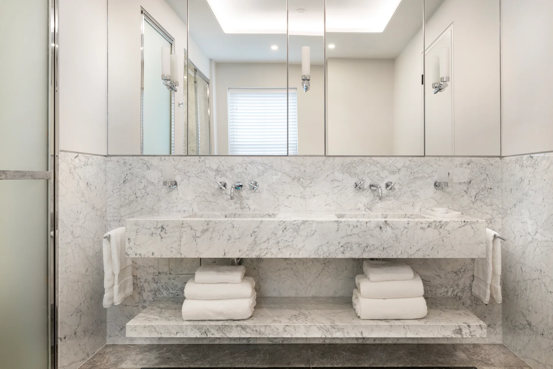 Modern bathroom featuring a marble sink, stacked towels, and large mirrors. Bright, elegant design with soft lighting.