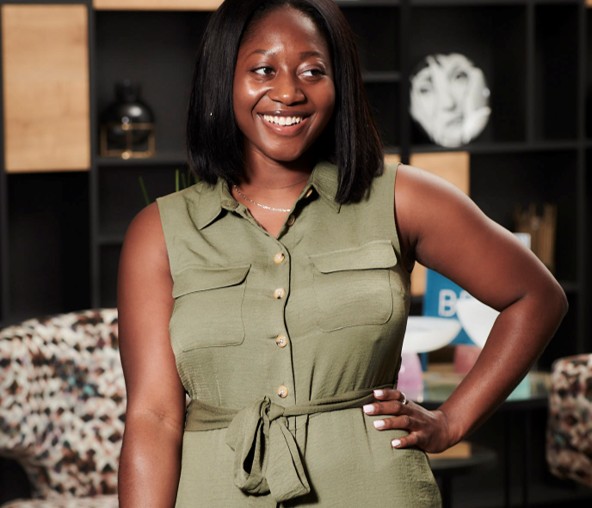 Smiling woman in a green dress with a waist tie, standing confidently in a modern studio setting.