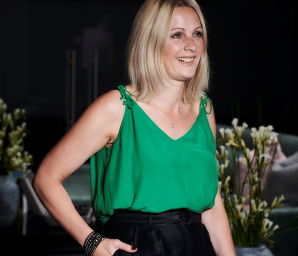 Woman with blonde hair wearing a green top and black pants, smiling, with flowers in the background.