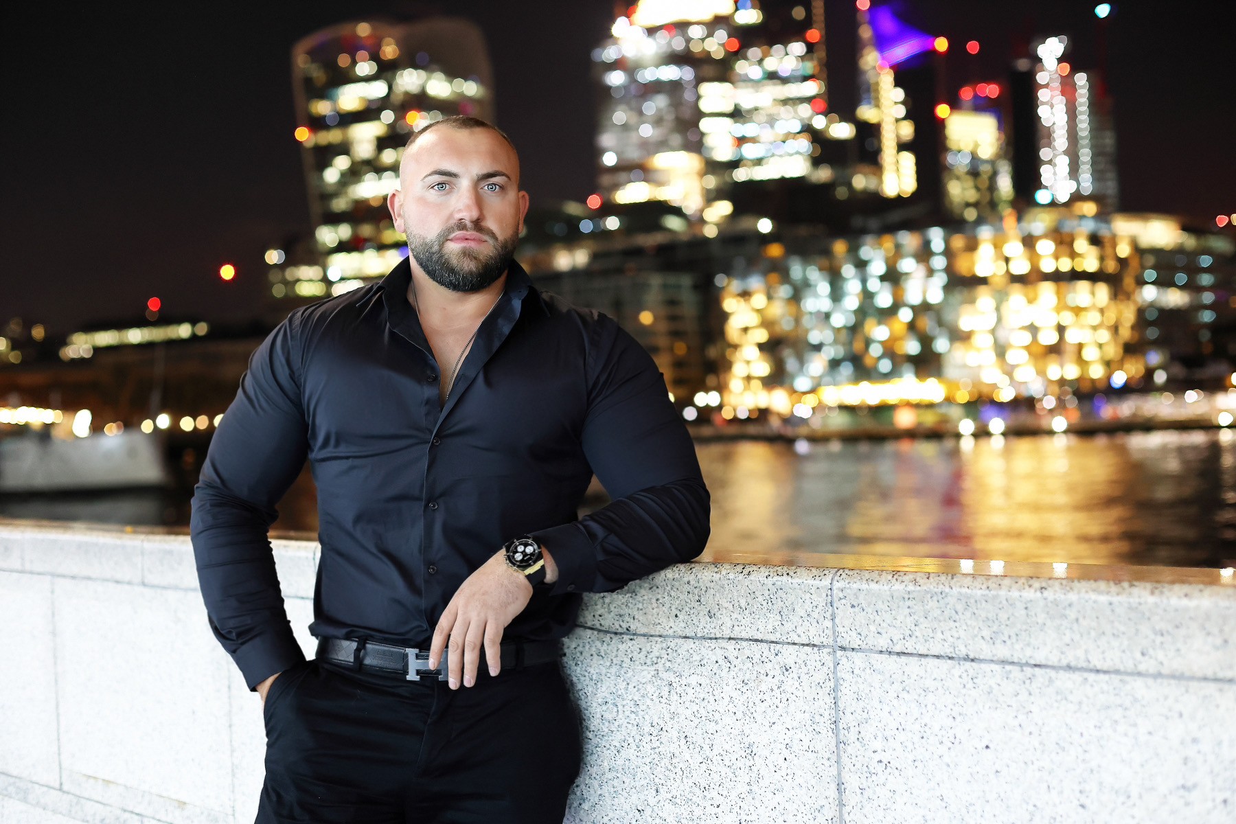 Man in a black shirt stands confidently by a riverside at night, illuminated city skyline in the background.