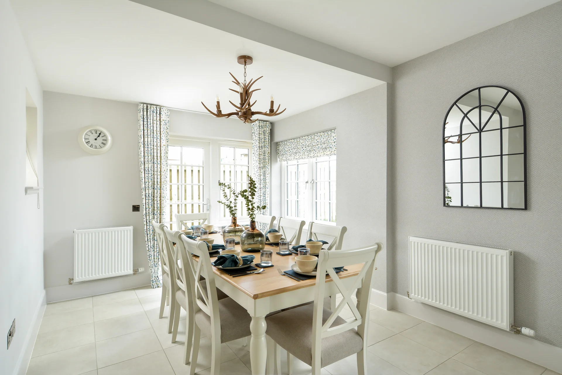 Bright dining room with wood table, white chairs, and large windows. Decorative chandelier and mirror enhance the space.