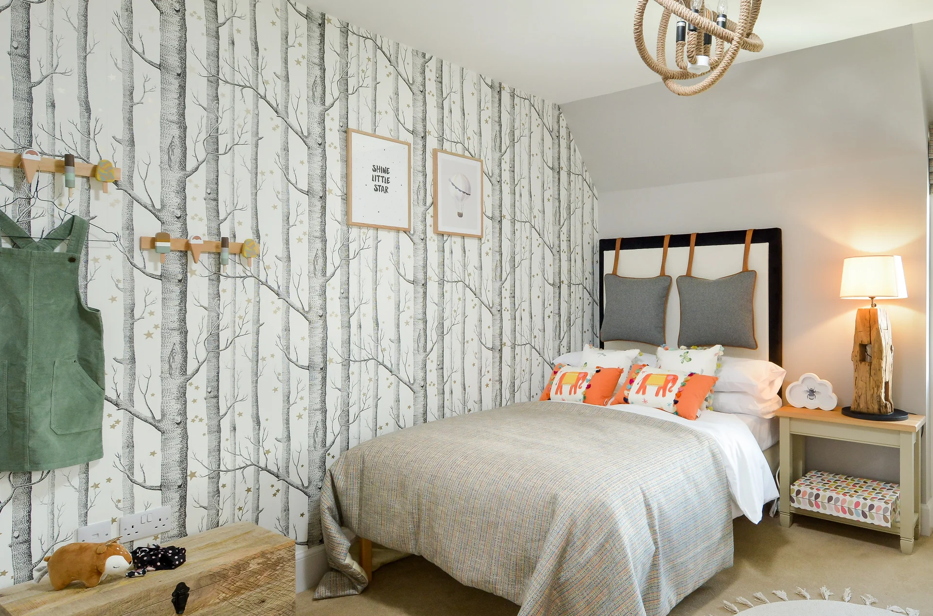 Cozy bedroom with a bed, decorative pillows, tree-patterned wallpaper, and a stylish lamp next to a nightstand.