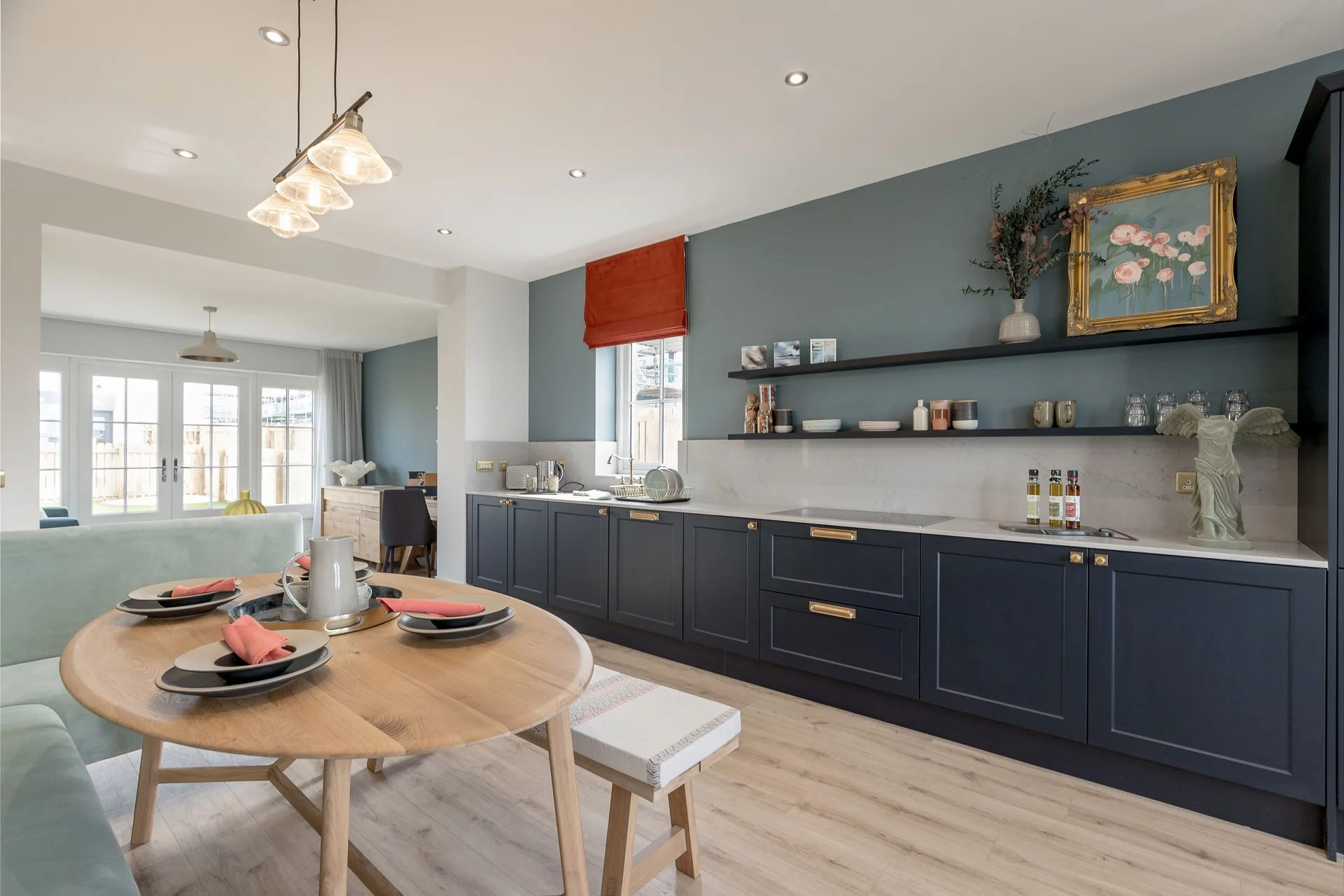Modern kitchen with blue cabinetry, wooden dining table set for four, and decorative wall art. Natural light from windows.