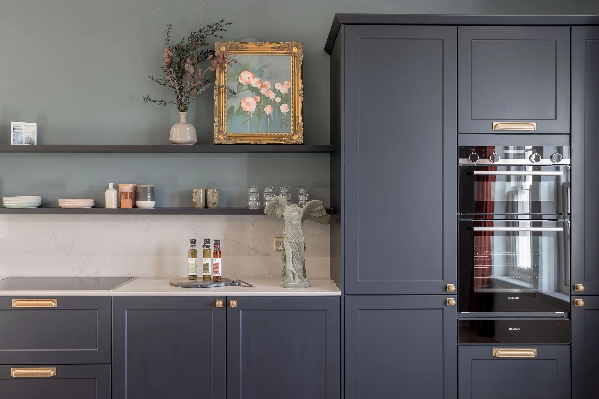 Modern kitchen with dark cabinetry, an oven, decorative shelf, and a floral painting on the wall.