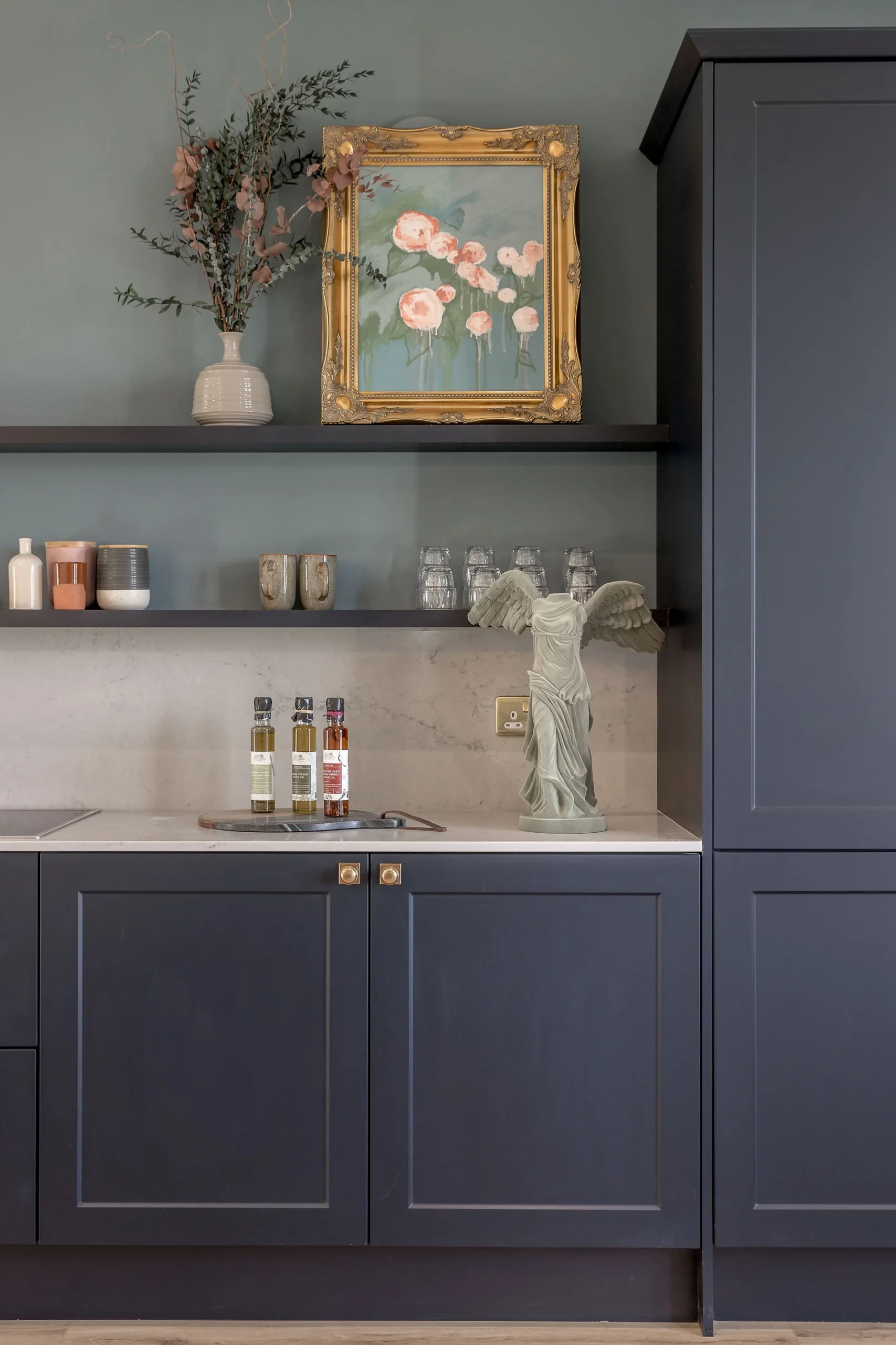 Modern kitchen with dark cabinetry, decorative shelf, vintage floral painting, and various kitchen items arranged neatly.