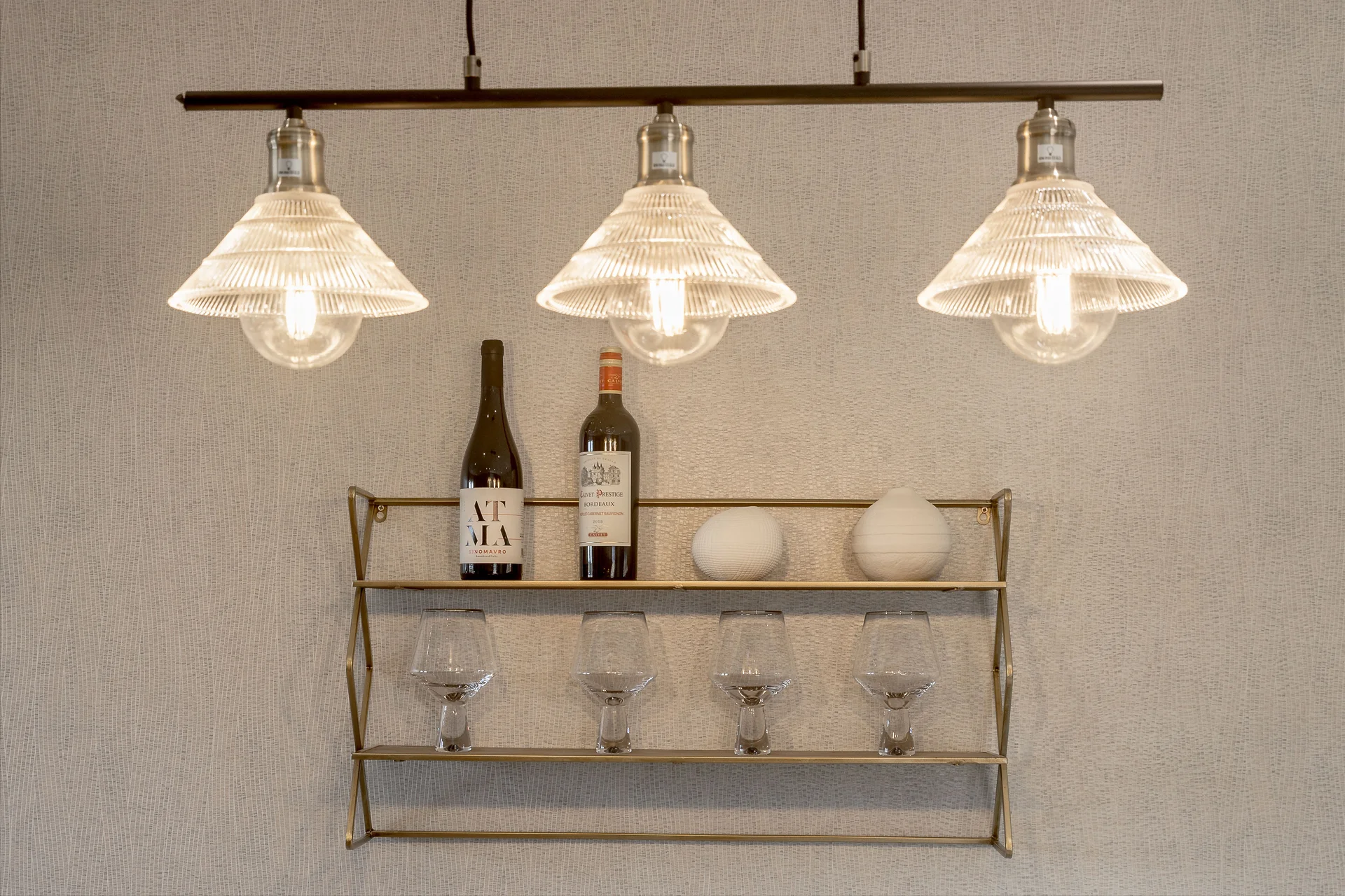 Three pendant lights hang above a shelf with wine bottles, glasses, and decorative items against a textured wall.
