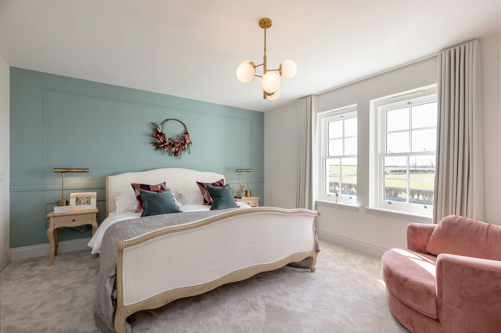 Brightly lit bedroom featuring a large bed, decorative wreath, two windows, and a pink accent chair.