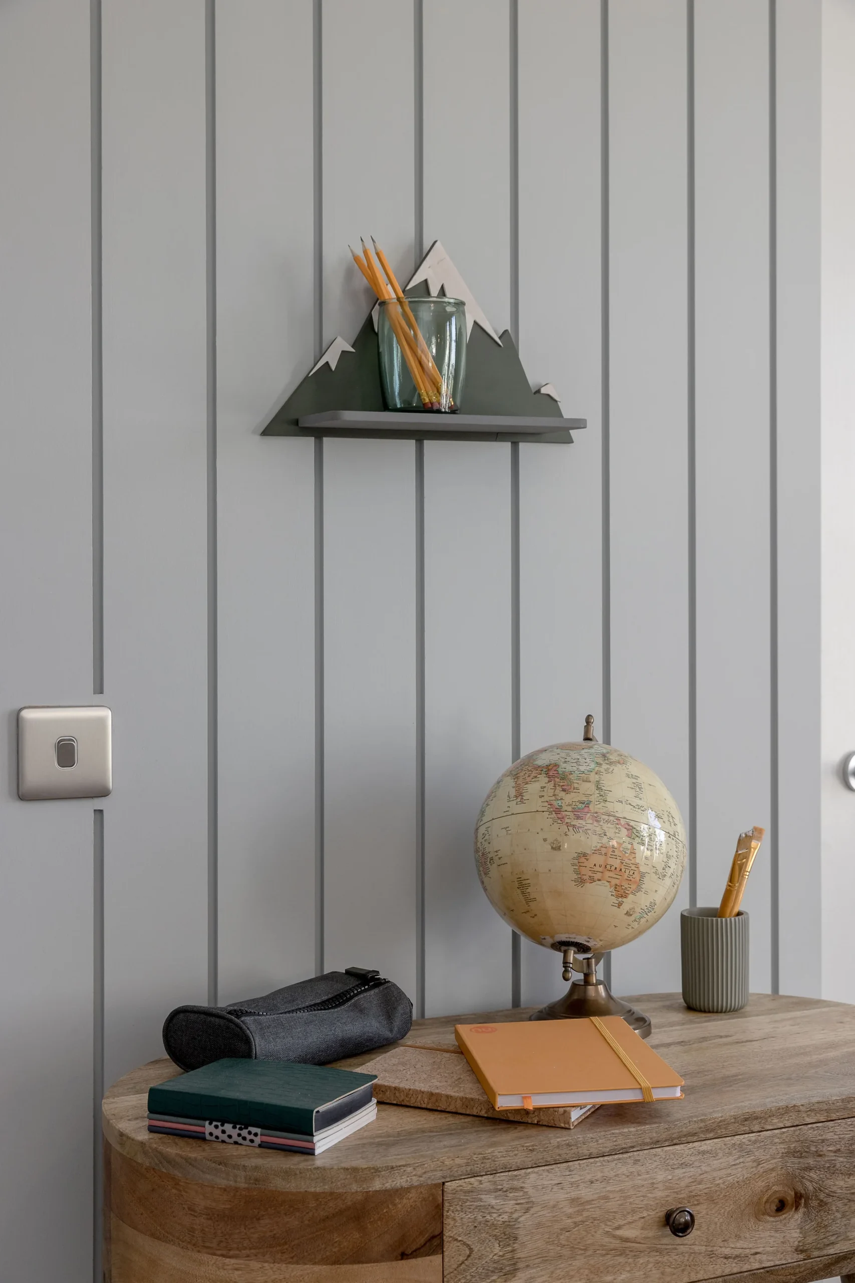 A wooden table with a globe, books, and a mountain-shaped shelf holding pencils against a gray wall.
