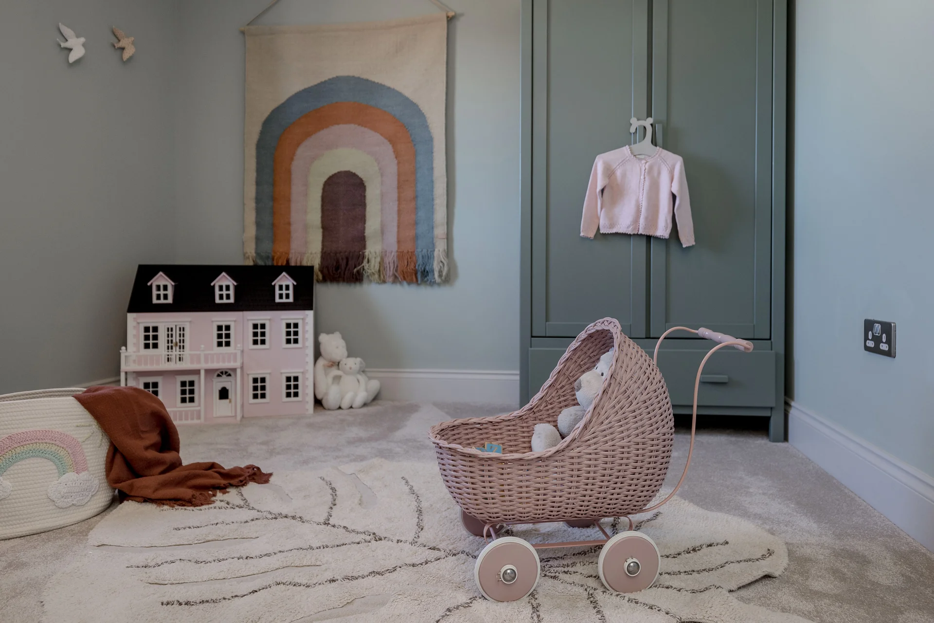 A stylish nursery featuring a woven doll stroller, a colorful rainbow wall hanging, and a small dollhouse.