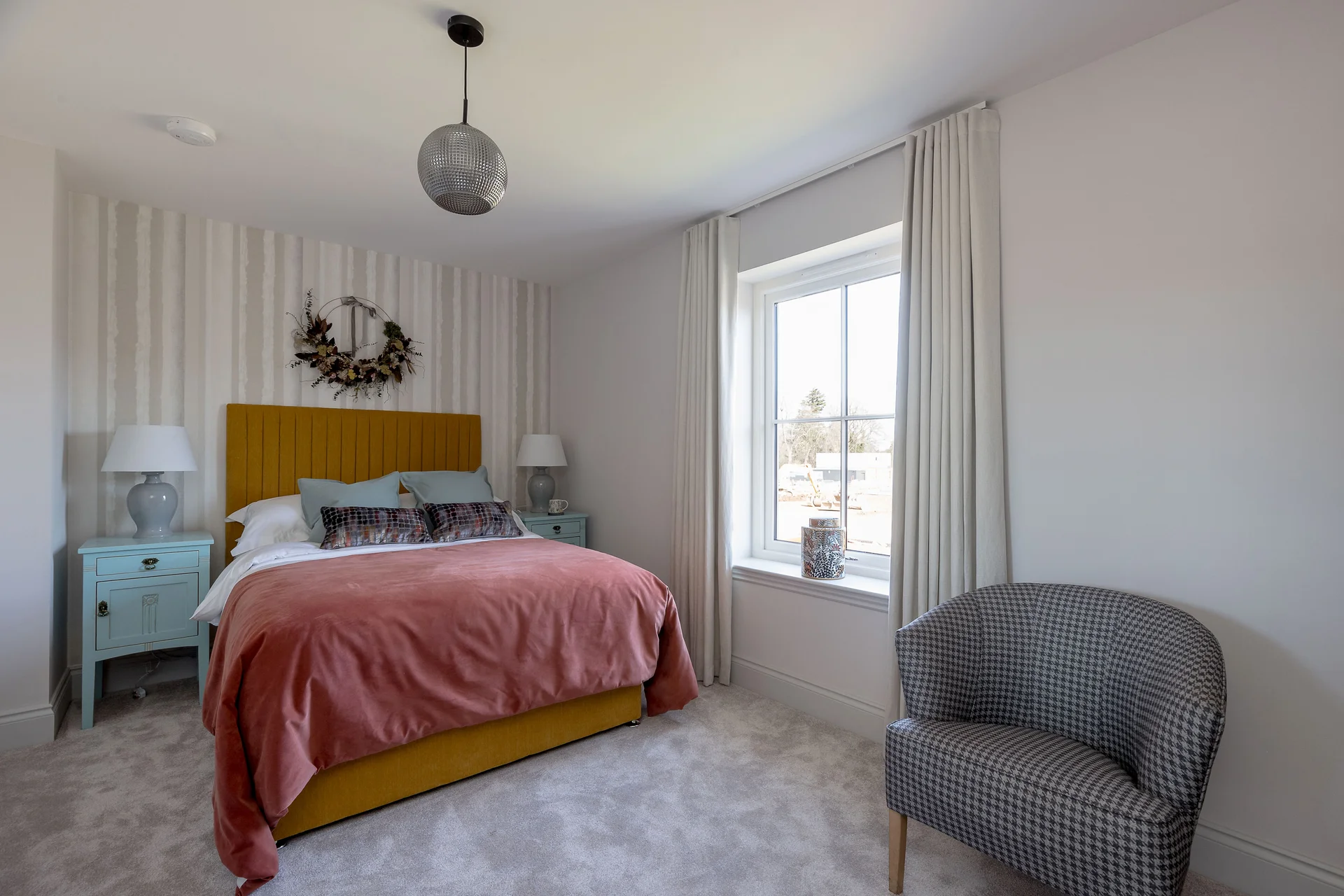 Cozy bedroom featuring a yellow bed with a pink throw, light blue nightstands, and a patterned chair near a bright window.