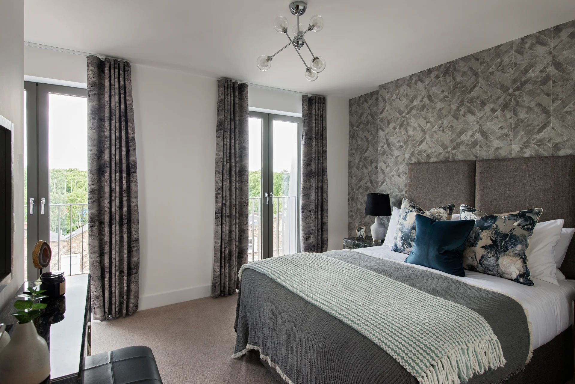 Modern bedroom with a double bed, decorative pillows, and large windows dressed in patterned curtains.
