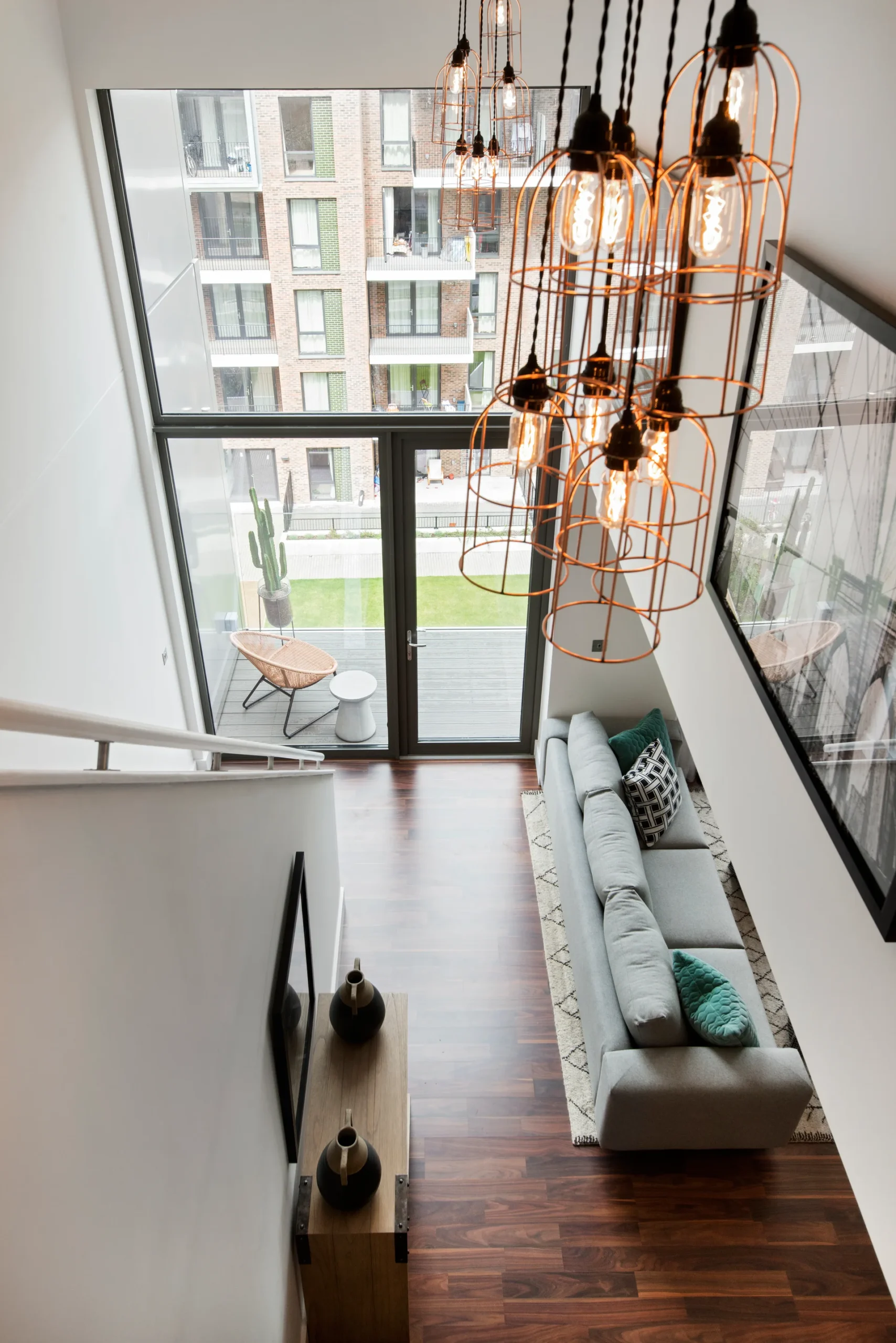 Modern living room with a gray sofa, wooden flooring, and pendant lights, featuring a large window overlooking an outdoor