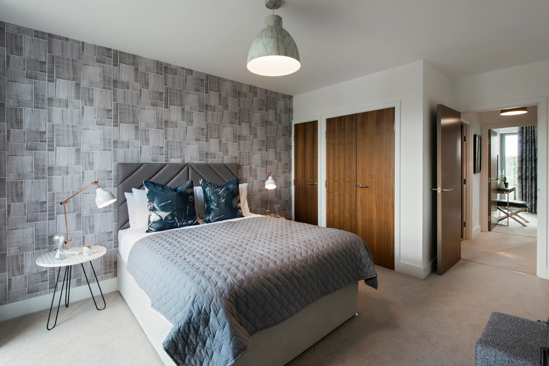 Modern bedroom featuring a double bed, decorative pillows, and stylish wallpaper, with doors leading to another room.