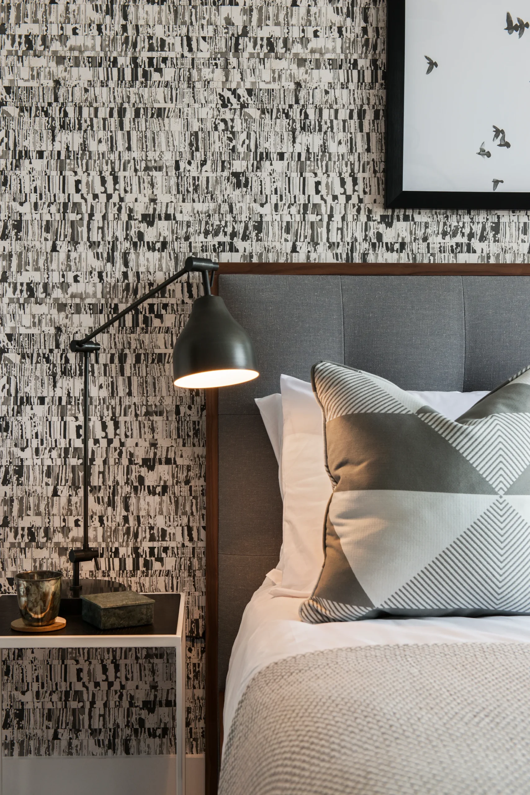 Modern bedroom featuring a gray headboard, geometric pillow, bedside lamp, and textured wallpaper.