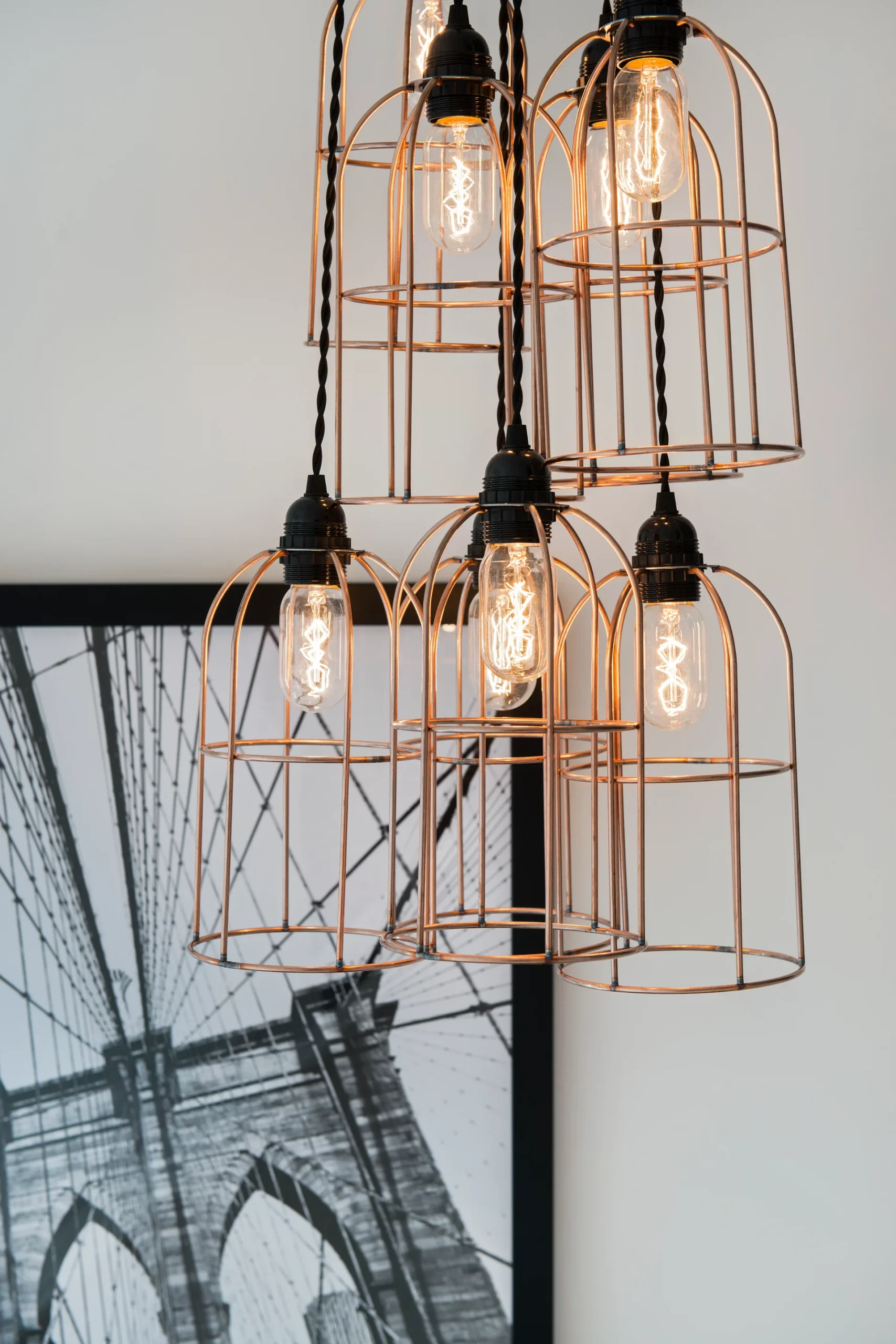 A pendant light with exposed bulbs enclosed in wire cages, hanging above a black-and-white photograph of a bridge.