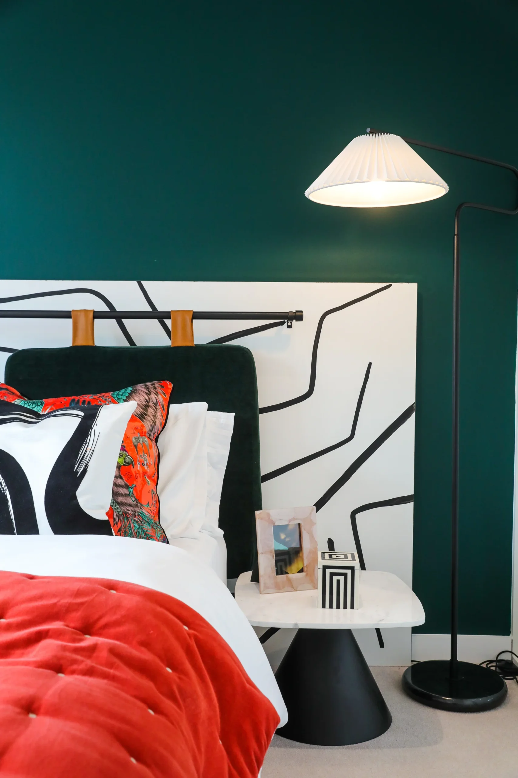 Stylish bedroom featuring a green wall, patterned headboard, red blanket, and modern lamp beside a minimalist nightstand.
