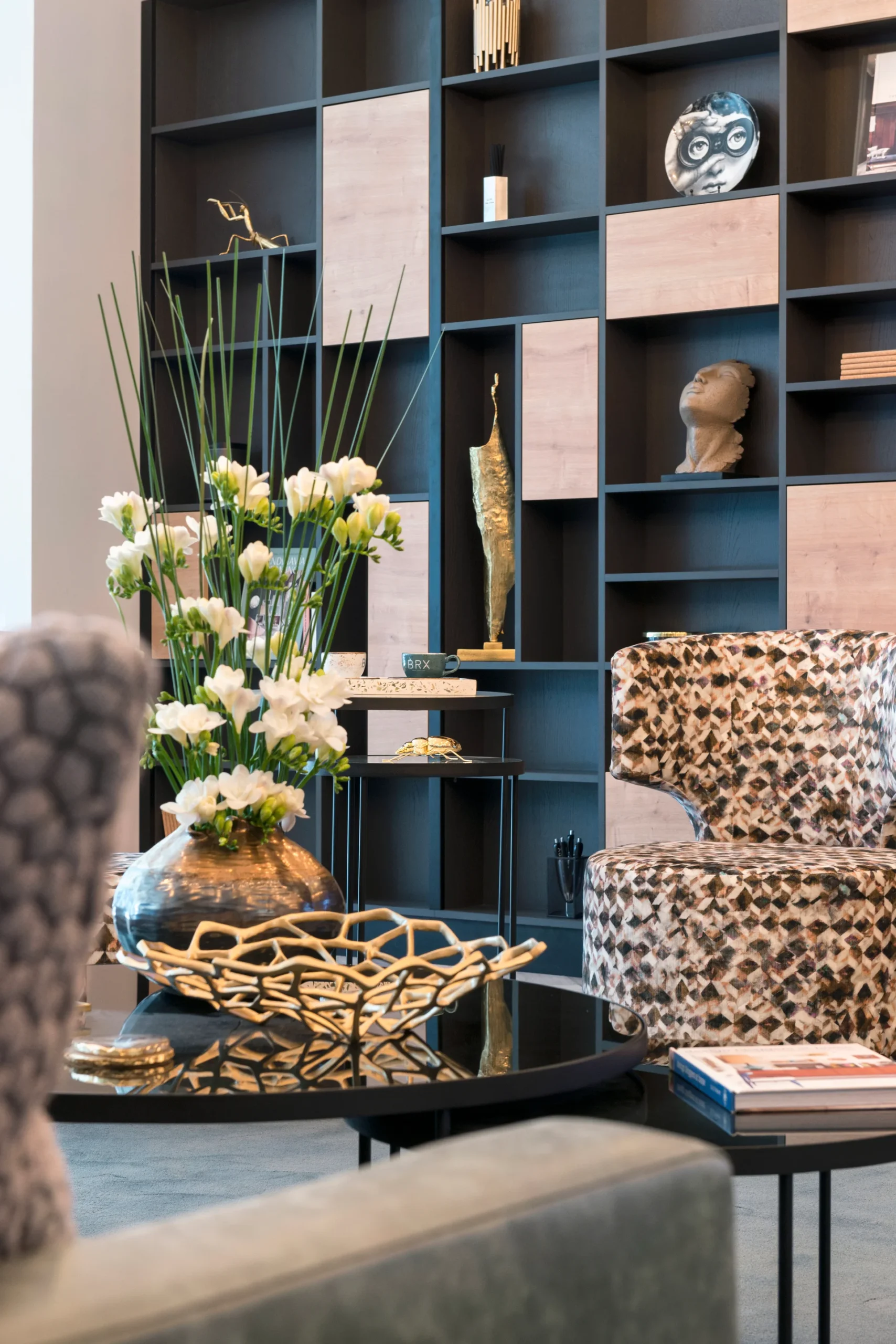 A stylish lounge area featuring a floral arrangement, decorative objects, and patterned chairs against a modern bookshelf.