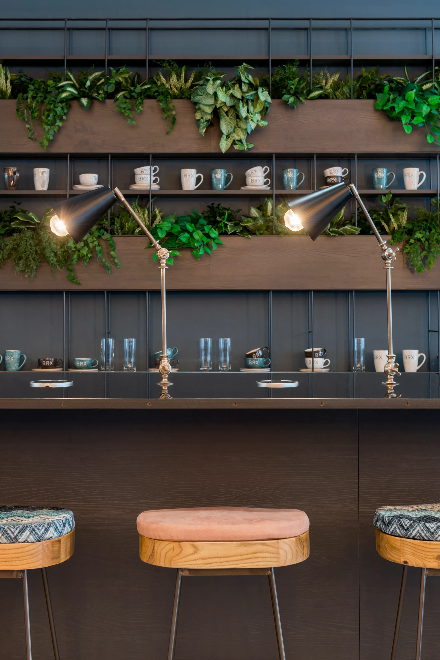 A modern coffee shop interior featuring a bar with two stools, surrounded by plants and decorative cups on shelving.