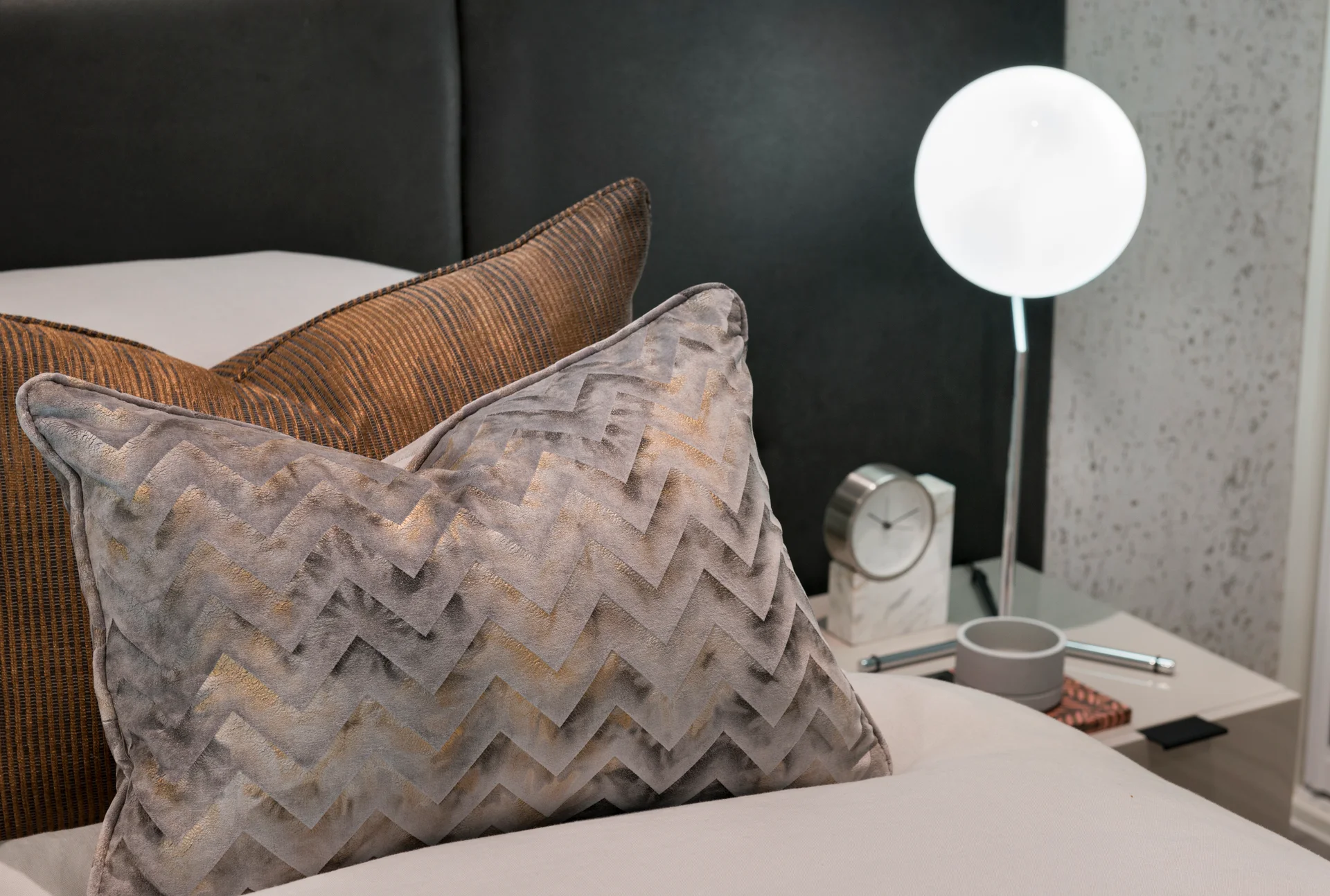 Gray and textured decorative pillows on a bed beside a modern lamp and bedside clock.
