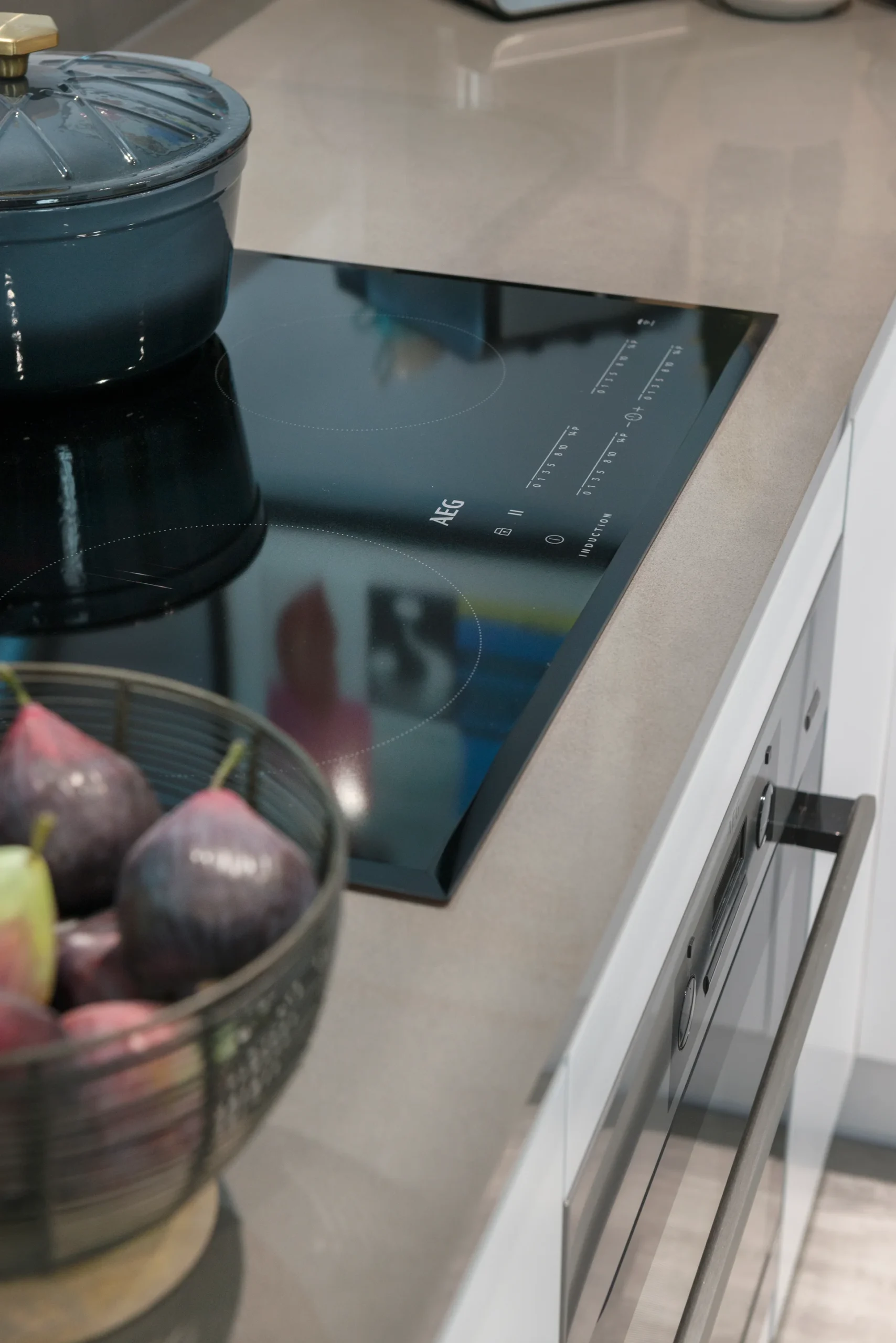 Modern kitchen countertop with a black induction stove, a blue pot, and a bowl of colorful fruits.